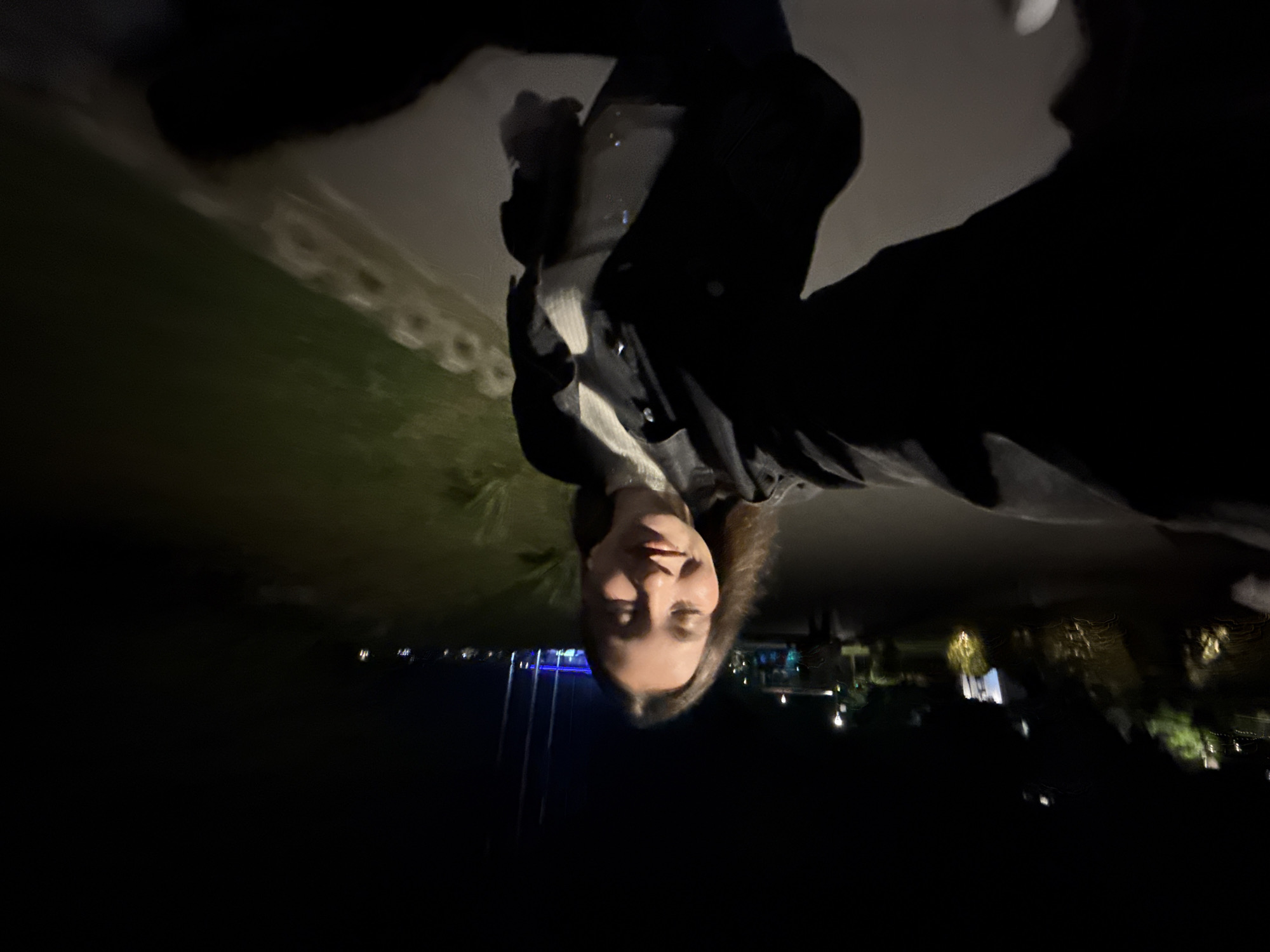 The image depicts a nighttime selfie taken by a female individual on a paved pathway in Sozopol, Bulgaria. The central subject is a young woman, positioned slightly off-center, holding the camera with her right arm extended, which is partially visible in the lower left foreground. She has shoulder-length brown hair and is wearing a dark jacket over a light-colored, ribbed top. Her eyes are closed, and she has a gentle smile, suggesting a relaxed or happy expression.
The scene is dimly lit by artificial light sources. To the left of the woman, a dark, vegetated incline is visible with multiple glowing, blurry light sources and structures, some exhibiting motion trails. A paved path extends behind her, flanked by a grassy area on the right. In the middle ground, streetlights illuminate portions of the path and sparse shrubbery. Further in the background, a structure with prominent blue lighting is visible, alongside three tall, dark poles, possibly flagpoles or antenna towers. The overall environment appears to be an outdoor pedestrian area or promenade. The foreground and bottom right show dark, blurry elements, likely parts of the subject's clothing or an object she is holding, contributing to the selfie perspective.