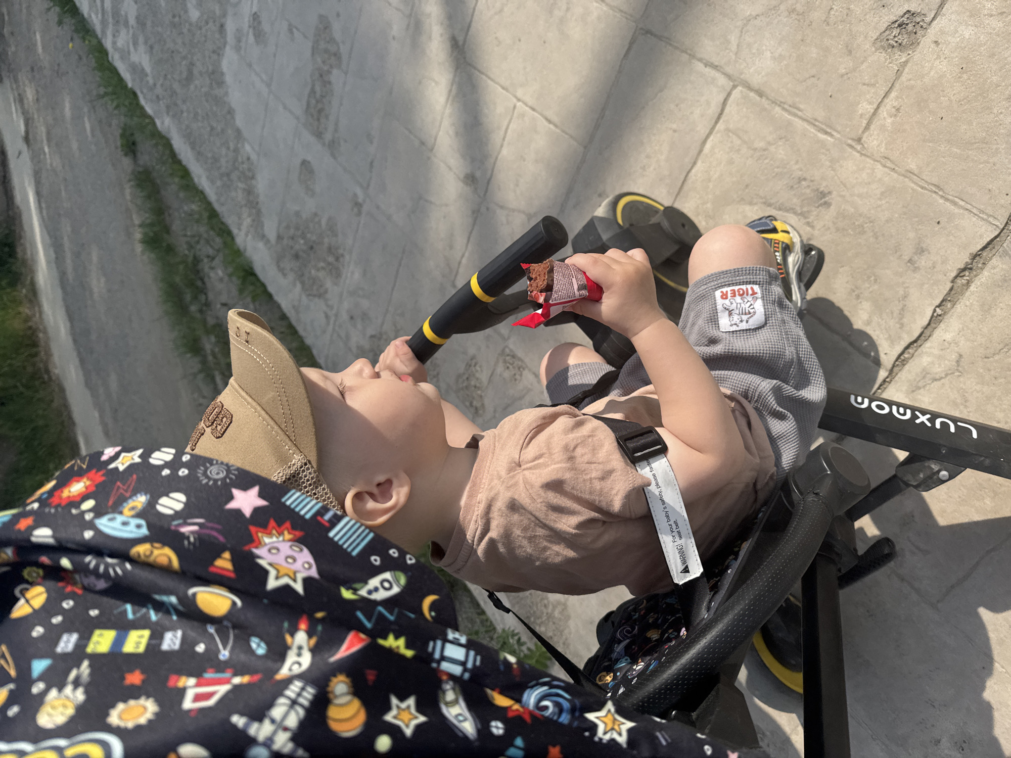 Captured from an elevated viewpoint, the scene depicts a young child seated in a black "LUXMON" stroller. The child, wearing a tan baseball cap and a muted pink t-shirt, is strapped into the stroller with a safety belt. His gray patterned shorts feature a "TIGER" logo. He holds a partly eaten chocolate brownie in its red wrapper. The stroller is positioned on a paved surface with stone-like patterns.
A portion of an adult's garment, patterned with cartoonish space-themed graphics, is visible in the lower left corner. The backdrop includes a concrete structure with hints of green foliage.
The image appears to be taken in daylight, possibly in Chaikovsky, Russia, though this cannot be verified definitively from the image alone. The focus is on the child and his interaction with the food, while the surrounding environment provides context.