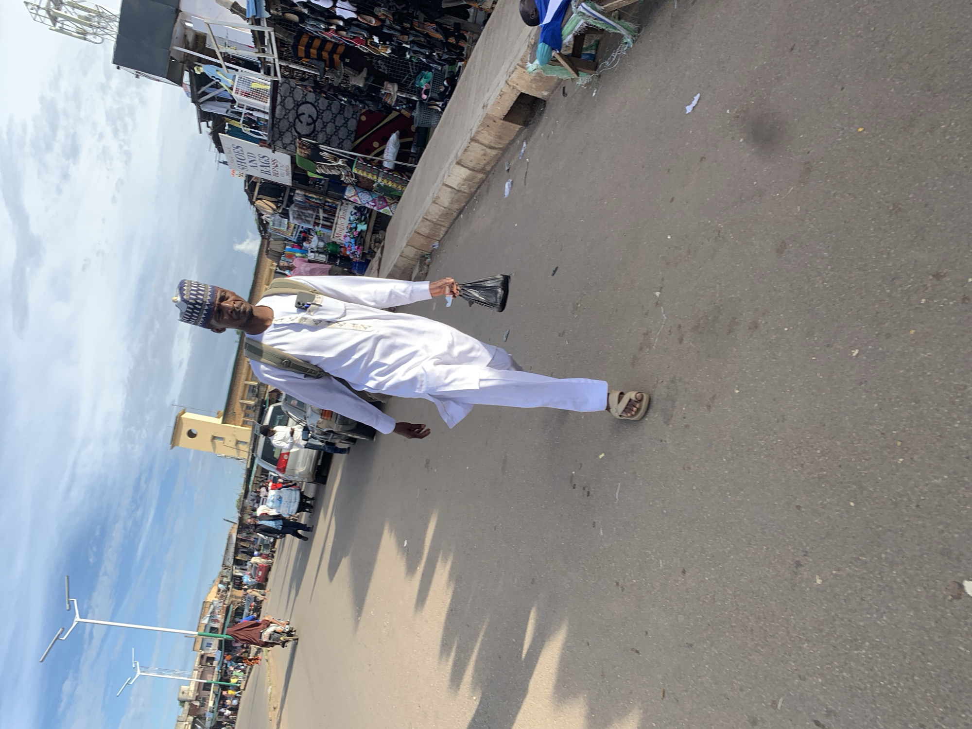 In the city of Jos, Nigeria, on a paved road, a man wearing a white tunic and trousers, and a patterned cap, walks towards the camera. He carries a black plastic bag in his right hand and a backpack on his shoulders. On his feet, he wears tan sandals. The road is lined with market stalls selling various goods, including shoes and bags. In the background, several vehicles are parked along the road, and streetlights stand tall. The sky above is partly cloudy.