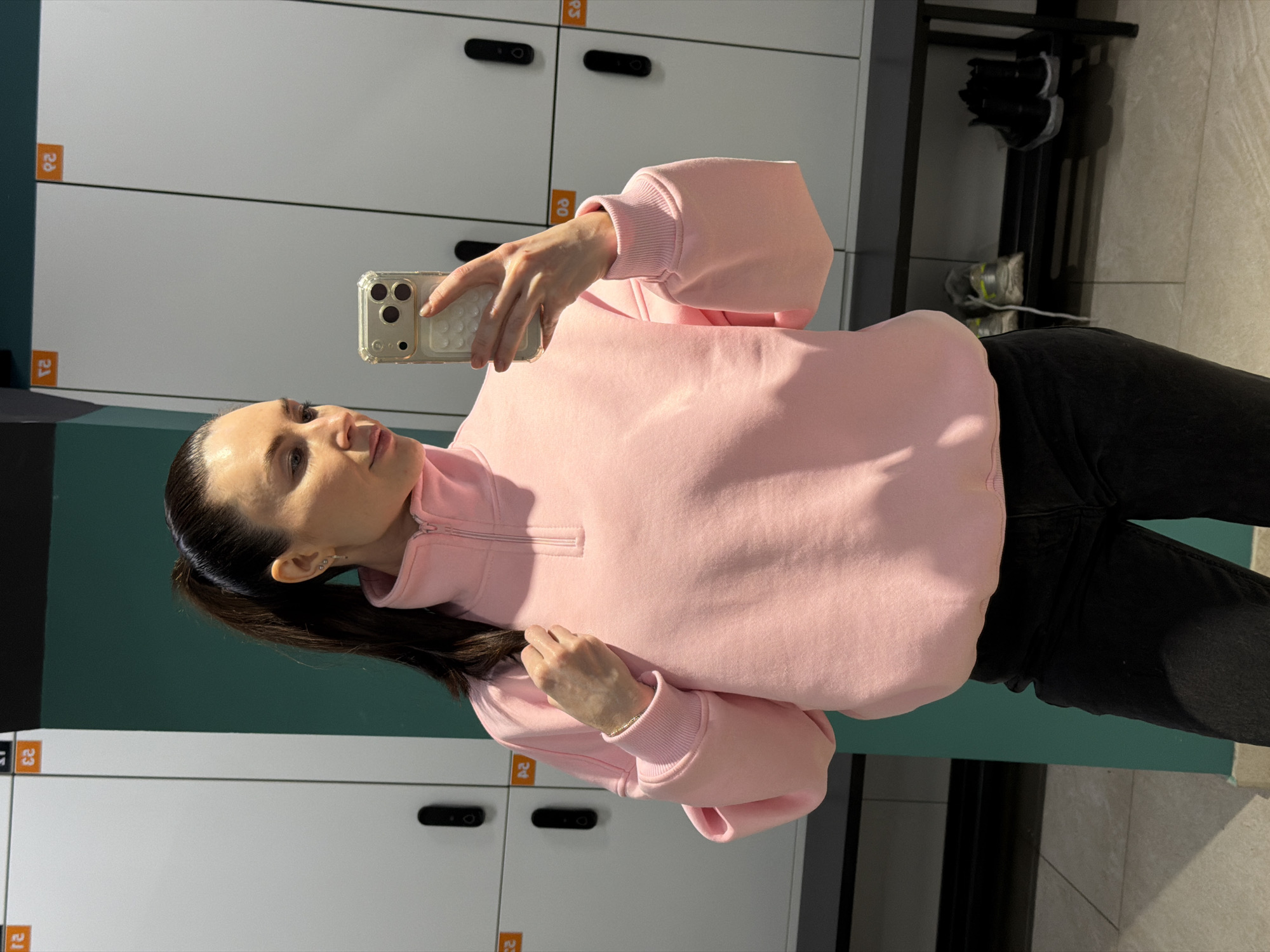 A woman is taking a selfie in a locker room. She is wearing a pink sweatshirt and black pants. Lockers with numbers (20-23, 03) are visible behind her. A black metal rack holding shoes can be seen in the background. The location is likely a gym or fitness center in Perm, Russia.