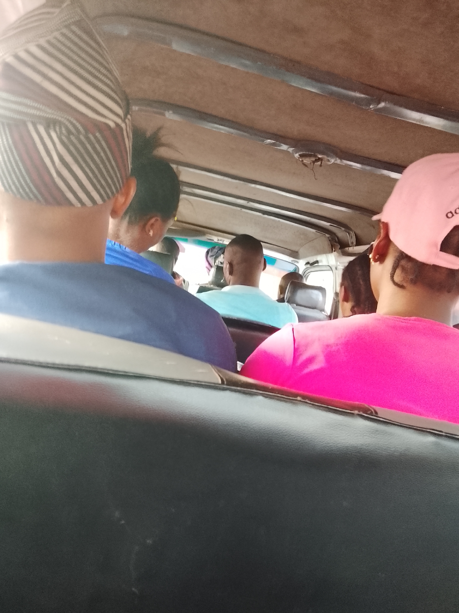 Captured from within a vehicle, the image shows a partial view of passengers and the interior. Five individuals are visible in the frame, their heads and upper backs appearing in a row. The person closest to the camera wears a hat with vertical white and maroon stripes, while the individual to their right wears a blue shirt. Further down, another passenger is dressed in a light blue shirt, and another has a pink shirt and matching pink baseball cap with white lettering. Black seats with a glossy finish are prominent in the foreground, and the vehicle's ceiling is a neutral tan color. Details visible on the ceiling include a metal support structure and a recessed light fixture. The image appears to have been taken in a public transportation setting.
