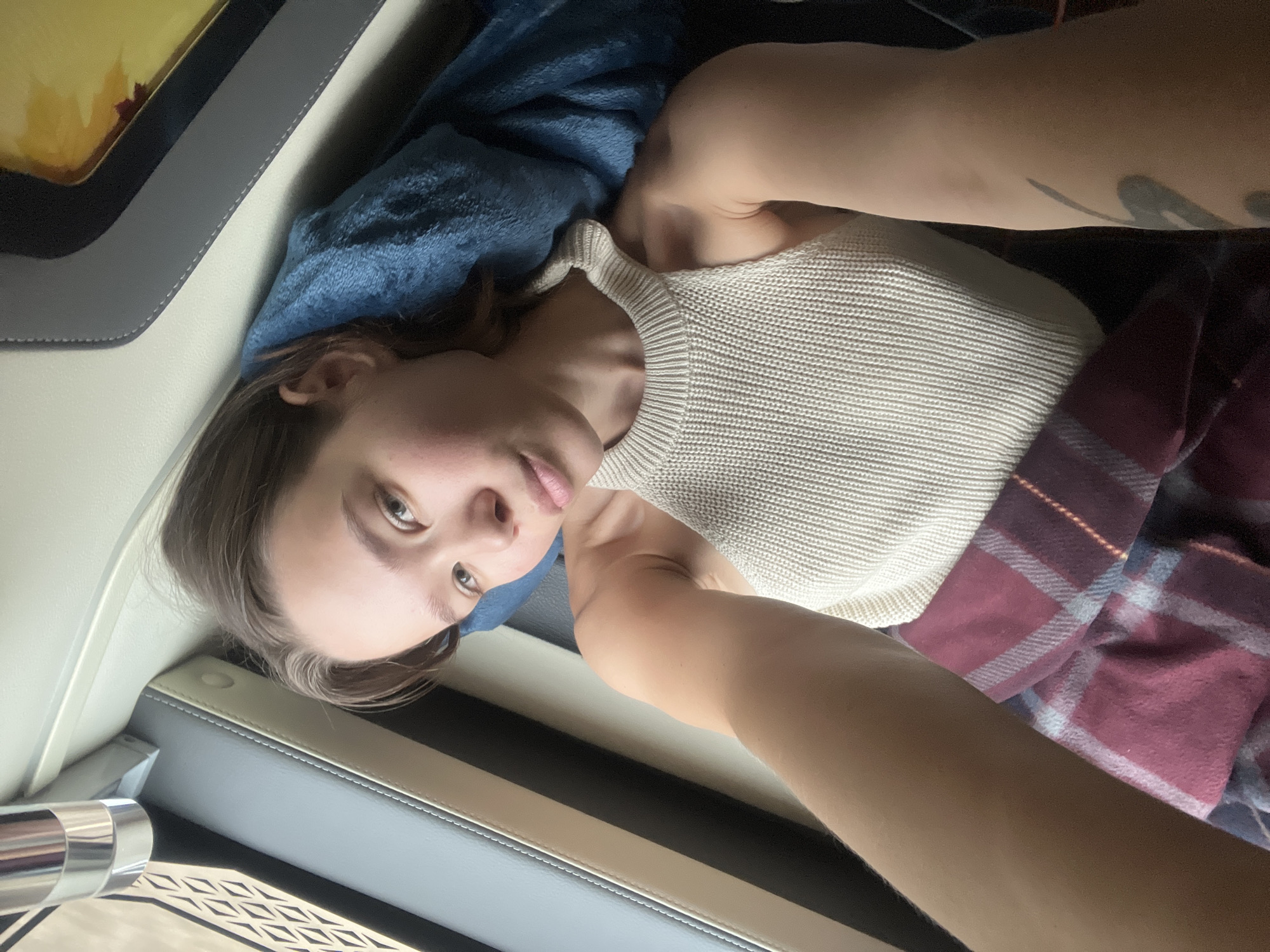 The image shows a young adult person, likely female, lying down and looking directly at the camera, suggesting a selfie. She is wearing a light-colored, possibly off-white, ribbed sleeveless top. Her brown hair is casually styled, and a tattoo is visible on her upper right arm.
She is resting with her head on a blue, textured blanket or pillow. A red and blue plaid blanket covers her lower body. The immediate surroundings indicate an interior space, likely a vehicle such as a car, bus, or van, given the upholstered seat-like structures, contrasting trim with visible stitching, and a metallic, cylindrical object on the left side of the frame, possibly part of the vehicle's console or a cup holder.
The person appears to be resting or relaxing during travel, consistent with the provided location context of Pờ Y, Vietnam. The lighting is soft and diffused, creating a calm atmosphere. There is no visible text in the image.
