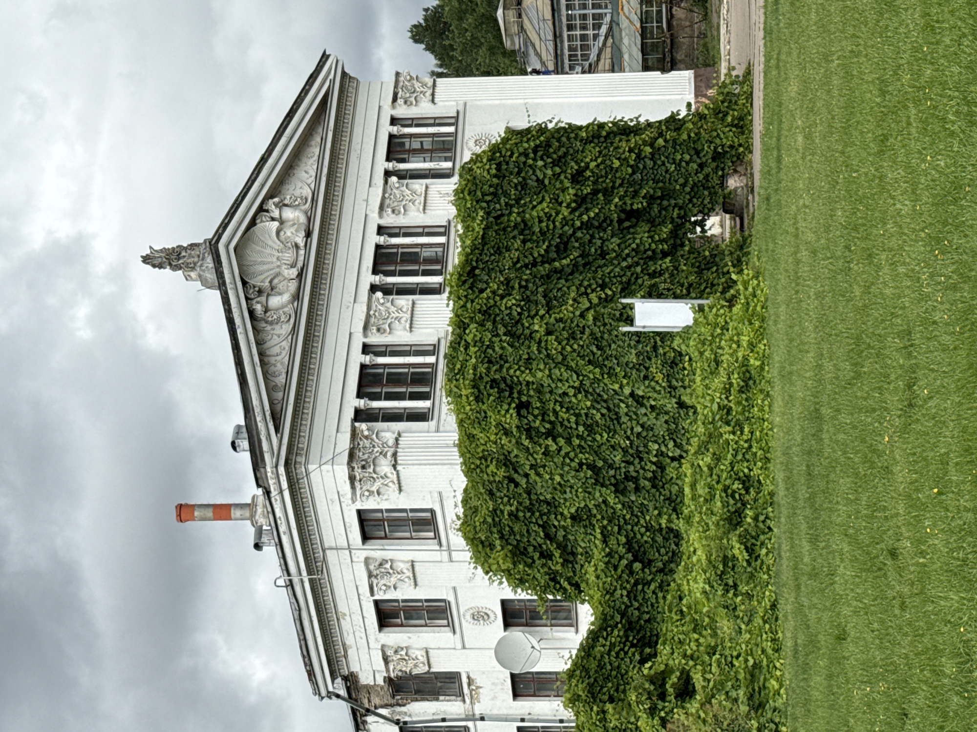 The image shows a white building with classical architectural elements, such as columns and pediments, partially covered by dense green foliage. A red and white striped chimney extends from the roof. The building has multiple windows with dark frames. In front of the building is a well-maintained green lawn. The sky is overcast with gray clouds. To the right of the main building, there are glimpses of greenhouse structures with glass panes. The scene appears to be an outdoor area within a city, possibly a park or botanical garden.
This building is the former main building of the Moscow State University Botanical Garden "Aptekarsky Ogorod".