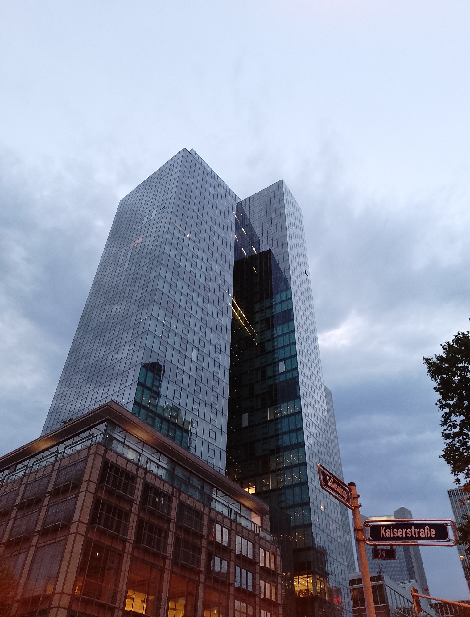 A low-angle, full shot captures a pair of modern, glass-clad skyscrapers and a lower, brick and glass building against a cloudy sky. The skyscrapers are tall and imposing, reflecting the overcast sky and the surrounding cityscape. The brick building in the foreground is a contrast to the sleekness of the skyscrapers, with its warm tones and grid-like window patterns. Two street signs are visible, one reading "Gallusanlage" and the other "Kaiserstrasse 29." The scene appears to be in an urban environment, possibly a business district, given the architecture. There are no people or animals visible in the frame. The overall mood is subdued, owing to the cloudy weather.