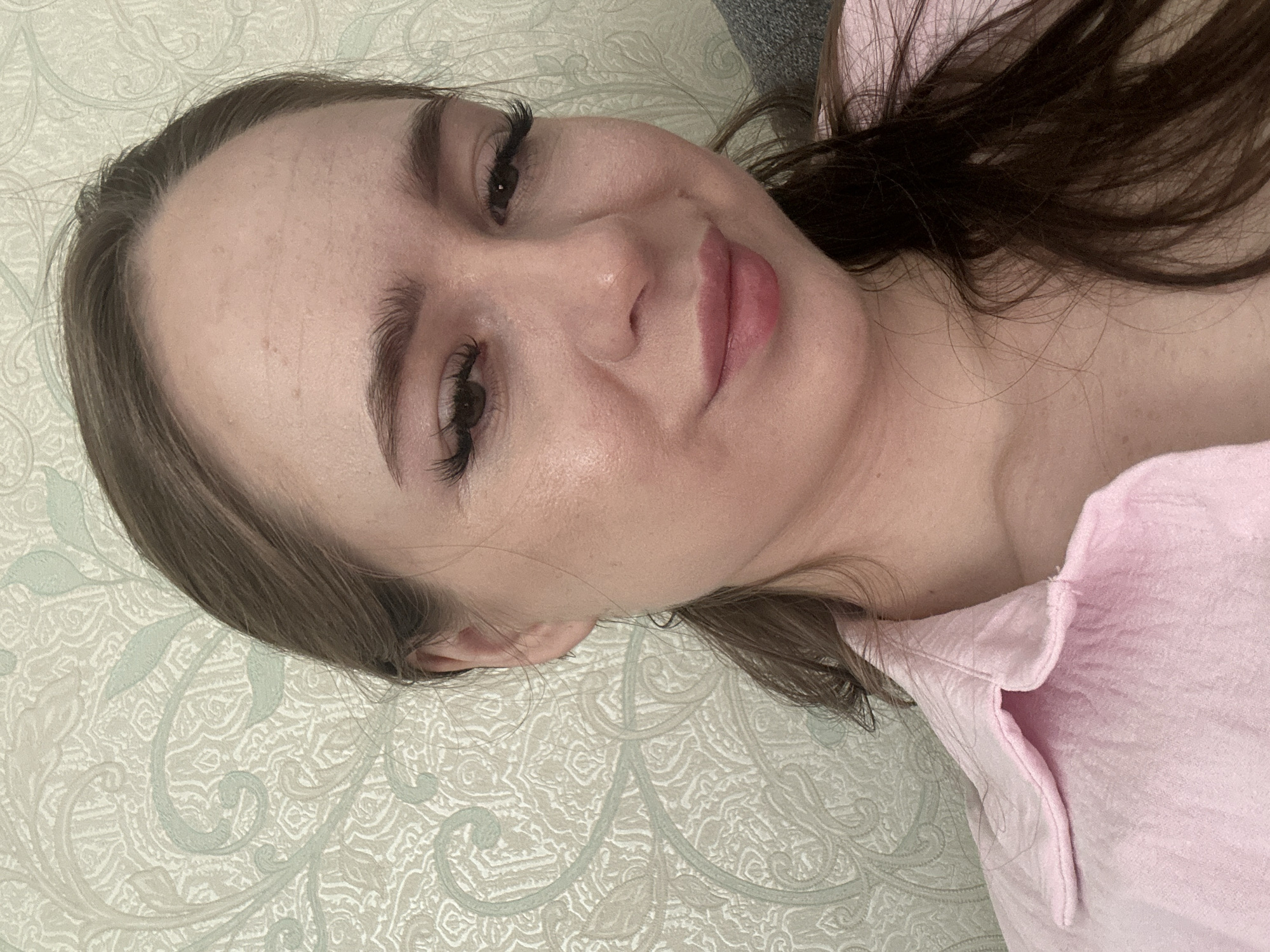 A medium close-up shot features a young woman with fair skin, brown hair tied back in a ponytail, and a pink collared shirt. Her makeup includes mascara, eyeliner, and pink lipstick. The background is a wall with a light-colored, ornate pattern. The lighting appears to be natural and soft, casting a gentle glow on her face. The image is set in an indoor environment, likely a home, in Unknown, Unknown.