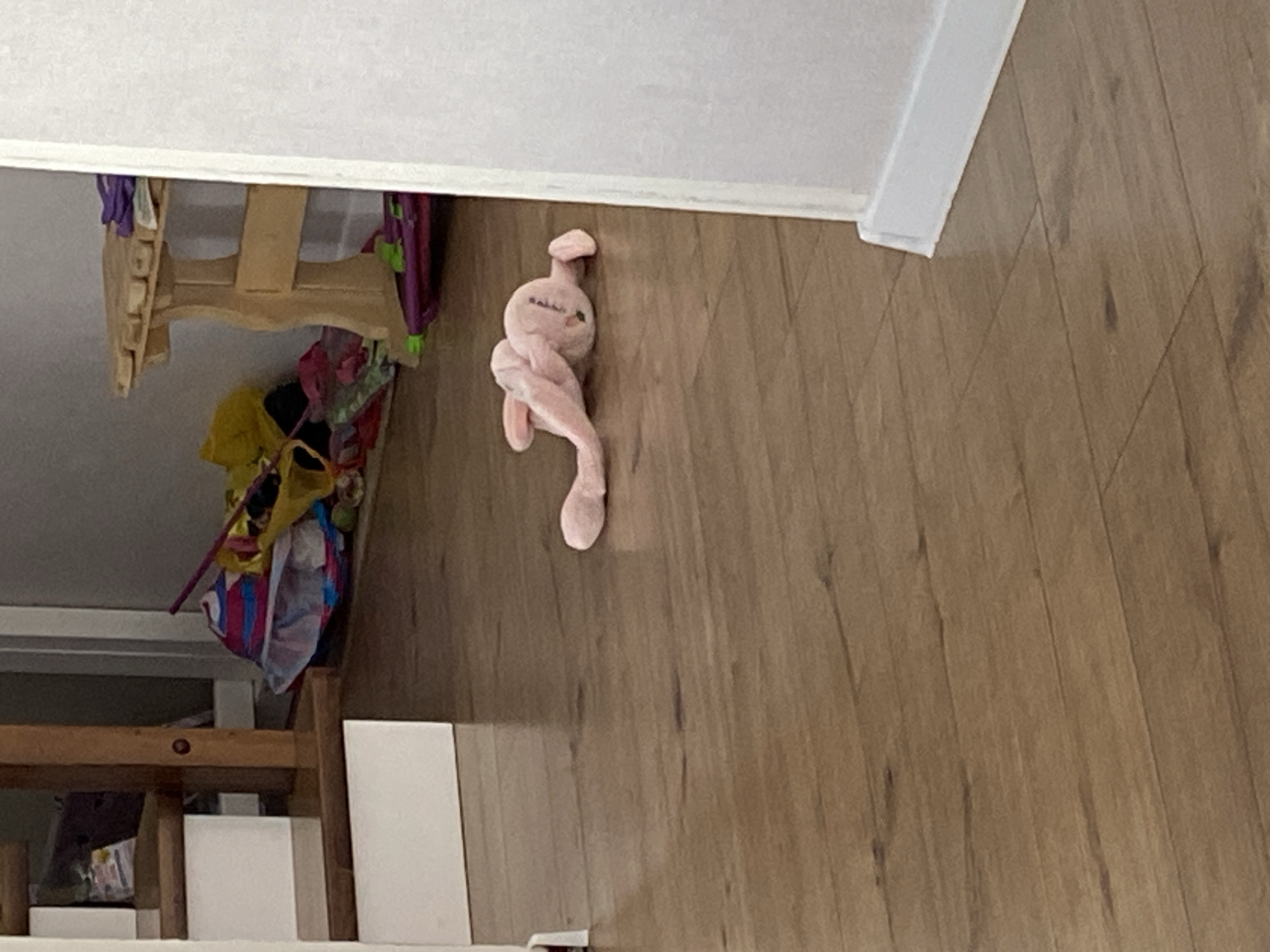 A stuffed pink bunny lies on its side on a wooden floor. The bunny is in the foreground of the image and is lying in the middle of the floor. The bunny is pink and has long ears. The bunny is lying on its side with its legs extended. In the background, there are stairs and a wooden table. There are also some toys and bags scattered on the floor. The lighting is dim, and the overall scene is slightly cluttered. The location is unknown.