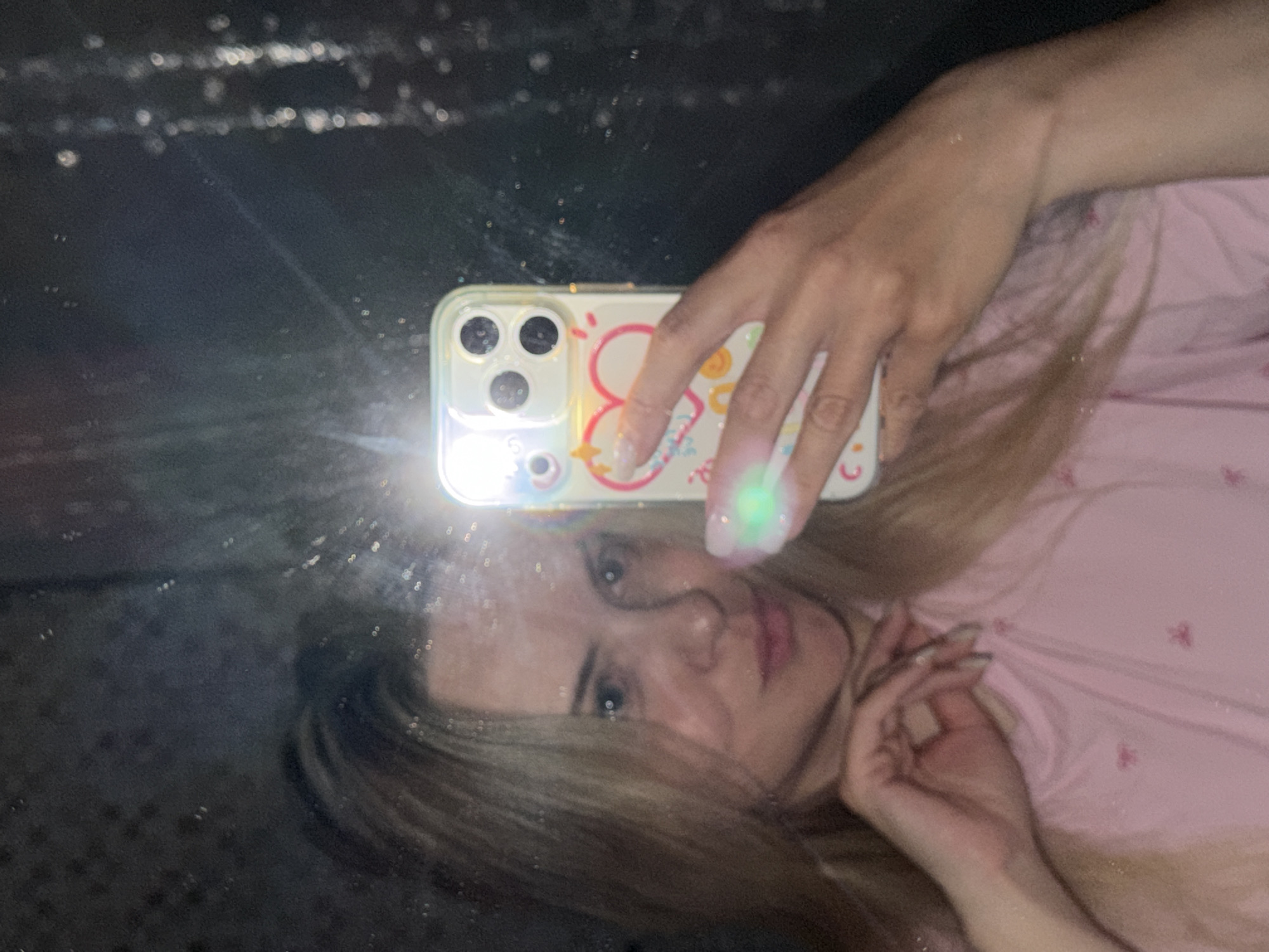 The image shows a woman taking a selfie in a mirror. She is holding a phone with a light-colored case decorated with hearts and other designs. The phone's camera flash is on, creating a bright spot of light. The woman has blonde hair and is wearing a pink top with a small pattern. The mirror reflects a dark background with some light reflections. The scene appears to be indoors. The image does not contain information indicating that the location is in Votkinsk, Russia.