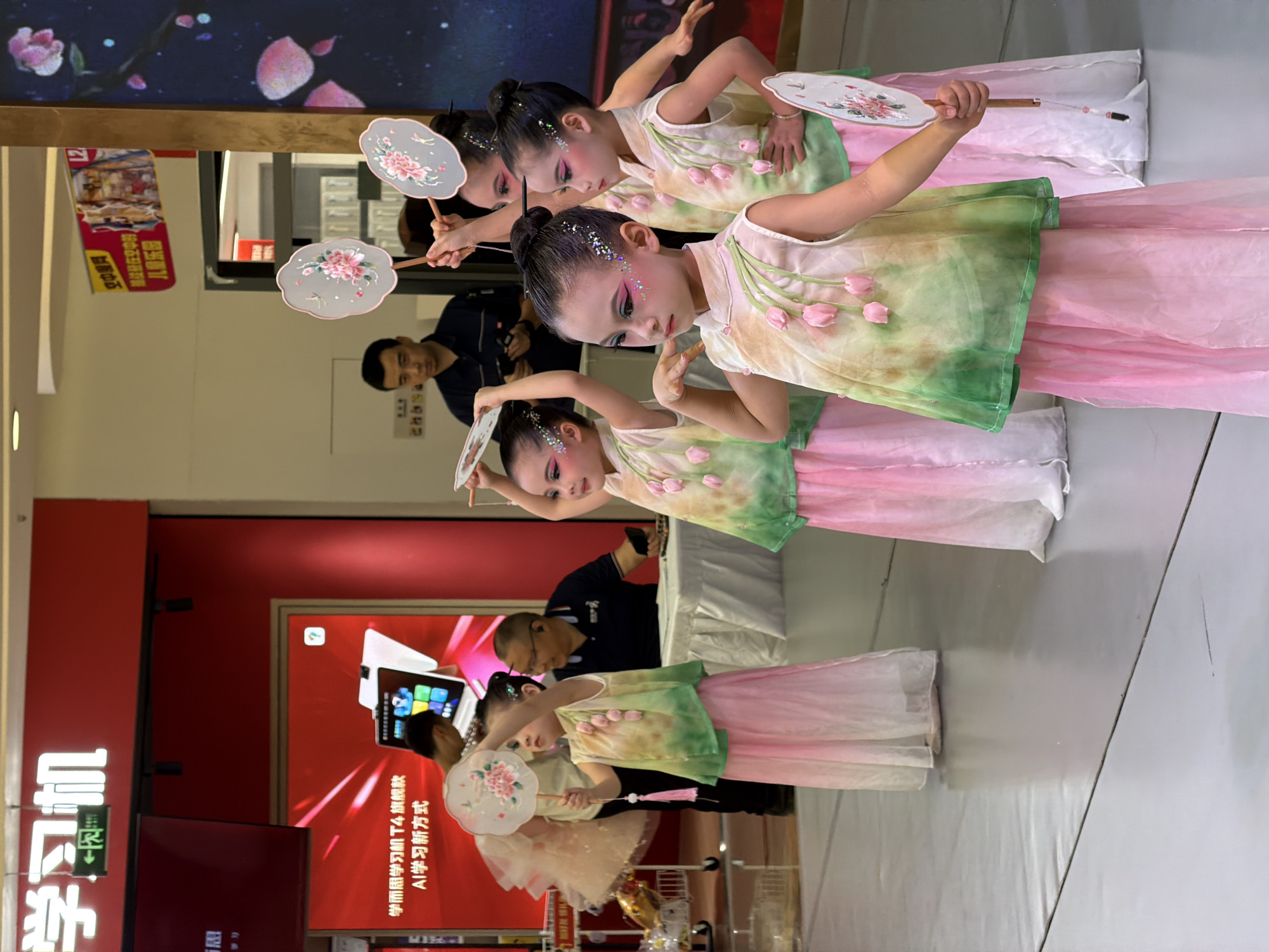 In Jiulongpo District, China, a group of young girls are performing a dance in what appears to be a commercial space, possibly a mall. They are dressed in traditional-inspired costumes featuring a gradient of pink and green, with floral embellishments and flowing skirts. Each girl holds a decorative fan, also adorned with floral patterns. Their faces are made up with theatrical makeup, including glitter on their eyelids and cheeks. In the background, several adults are visible. One man in a dark shirt is standing and looking towards the performers, while another man, wearing glasses and a blue shirt, sits at a table and looks at his phone. A large digital display screen is mounted on the red wall behind the man sitting at the table, advertising a "learning machine." The floor is a light gray surface, and the overall lighting suggests an indoor environment.