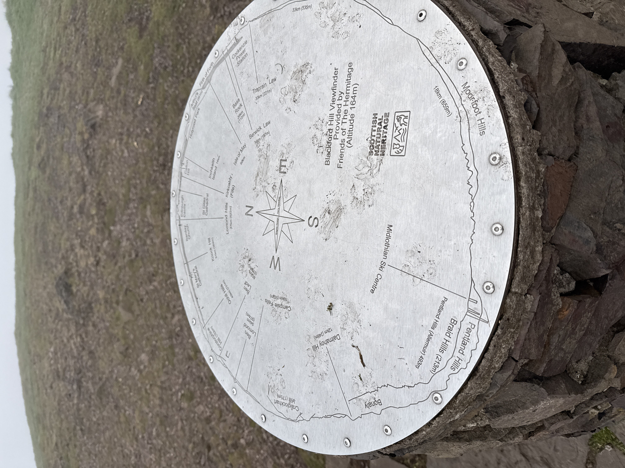 A circular, silver-colored metal viewpoint indicator, also known as a viewfinder, is prominently featured, secured to a rough, dark stone base with multiple rivets. The disc is engraved with a compass rose at its center, clearly marking North, East, South, and West. Radial lines extend from the compass, pointing towards various geographical landmarks.
The viewfinder is titled "Blackford Hill Viewfinder Provided by Friends of The Hermitage (Altitude 164m)," indicating its specific location and purpose. It also displays the "SCOTTISH NATURAL HERITAGE" logo. The surface of the disc shows signs of use and weather exposure, with dirt and smudges visible.
Numerous Scottish landmarks are identified, each with its distance from Blackford Hill and, in most cases, its altitude. These include:
- To the east: Traprain Law (33km, 221m), Berwick Law (34km, 187m), Bass Rock (38km, 112m), Isle of May (50km), Cockenzie Power Station, Firth of Forth.
- To the north: Arthur's Seat (251m), Holyrood Park, Edinburgh Castle, St Giles Cathedral, Calton Hill, Inchkeith Island (14km), Kirkcaldy (Fife), Lomond Hills (37km, 522m).
- To the west: Knock Hill (31km, 364m), Campsie Fells (59km, 570m), Ben Lomond (94km, 974m), Ben Ledi (80km, 879m), Ochil Hills (46km, 721m), Corstorphine Hill (162m), Craiglockhart Hill (175m), Dalmahoy Hill (12km, 246m).
- To the south: Bonaly, Midlothian Ski Centre, Pentland Hills (Allermuir 493m), Braid Hills (213m), and Moorfoot Hills (18km, 659m).
The background reveals a hazy, possibly misty or overcast outdoor environment, consistent with a hilltop vantage point, showing indistinct green and brown terrain stretching into the distance. The image captures a functional outdoor informational display on a cloudy day.