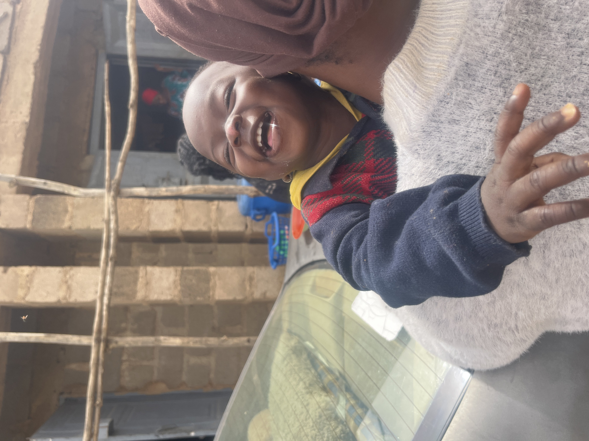 A child with dark skin and a missing front tooth is held up by a person in a beige sweater, smiling and waving. The child is wearing a dark blue sweater with a red and yellow plaid pattern on the front. A second person is partially visible in the background. The location appears to be Jos, Nigeria. A building under construction with exposed brick and scaffolding is visible behind the child and people. A car is partially visible at the bottom of the frame.