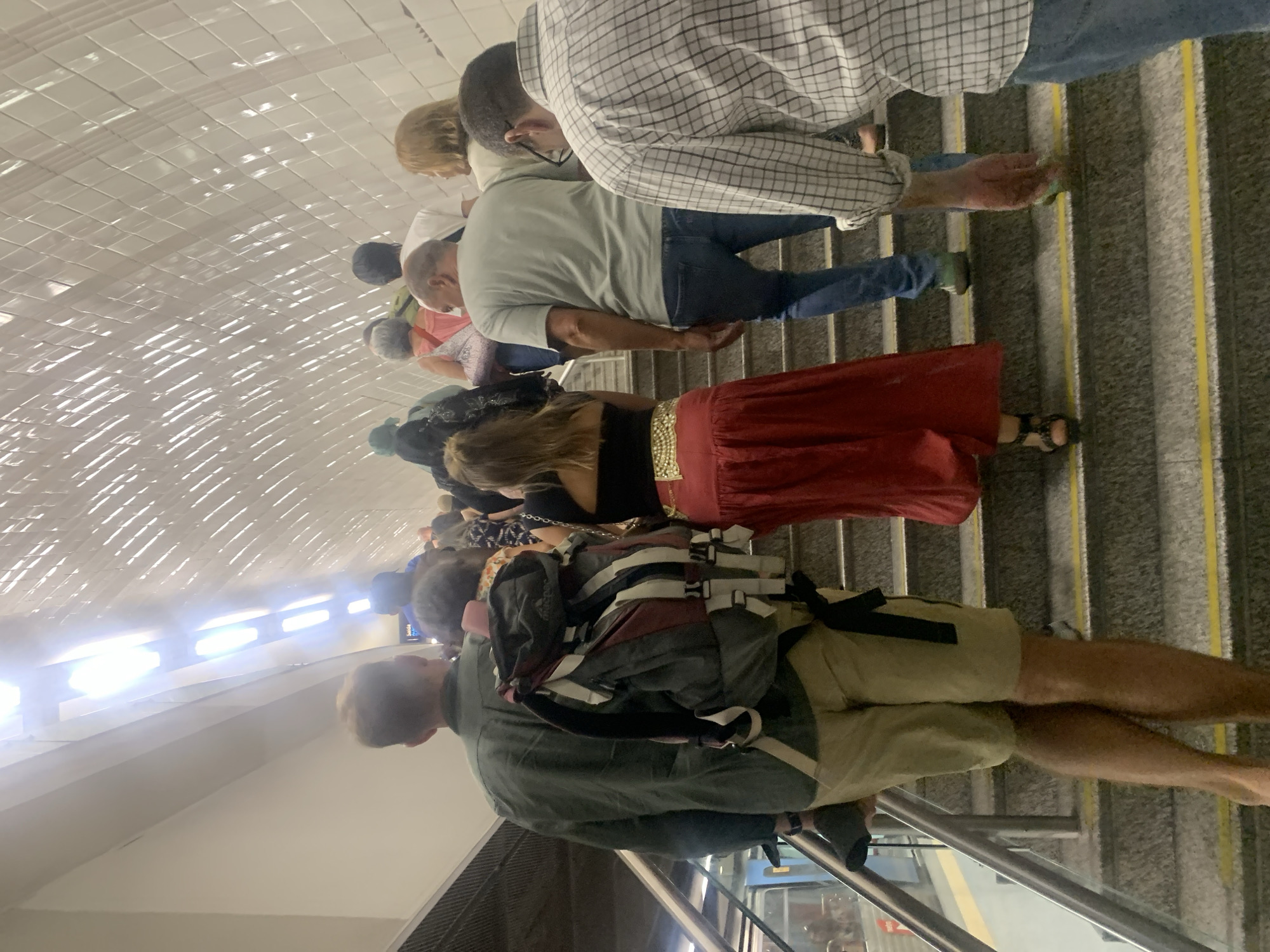 This media file shows a group of people on an escalator in what appears to be a subway station in Lisbon, Portugal. The subjects are diverse in age and gender, and are dressed in casual clothing. Some are carrying backpacks, suggesting they might be tourists or commuters.
The scene is set inside a modern subway station with white tiled walls and bright fluorescent lighting, characteristic of public transportation infrastructure. The escalator steps are grey with yellow safety lines. The overall atmosphere seems neutral, with no overt emotional displays. It is difficult to determine the time of day or weather conditions from the image.
The people are in various stages of ascending the escalator. Some are looking forward, others are glancing at their phones or engaged in conversation. One individual in the foreground is wearing a green jacket and khaki shorts, with a large backpack. Further up the escalator, a person in a red skirt and black top stands out. Another person in a checkered shirt is visible near the top of the frame.
No visible text is present in the image. The context of Lisbon, Portugal, suggests this might be a scene from the city's metro system.