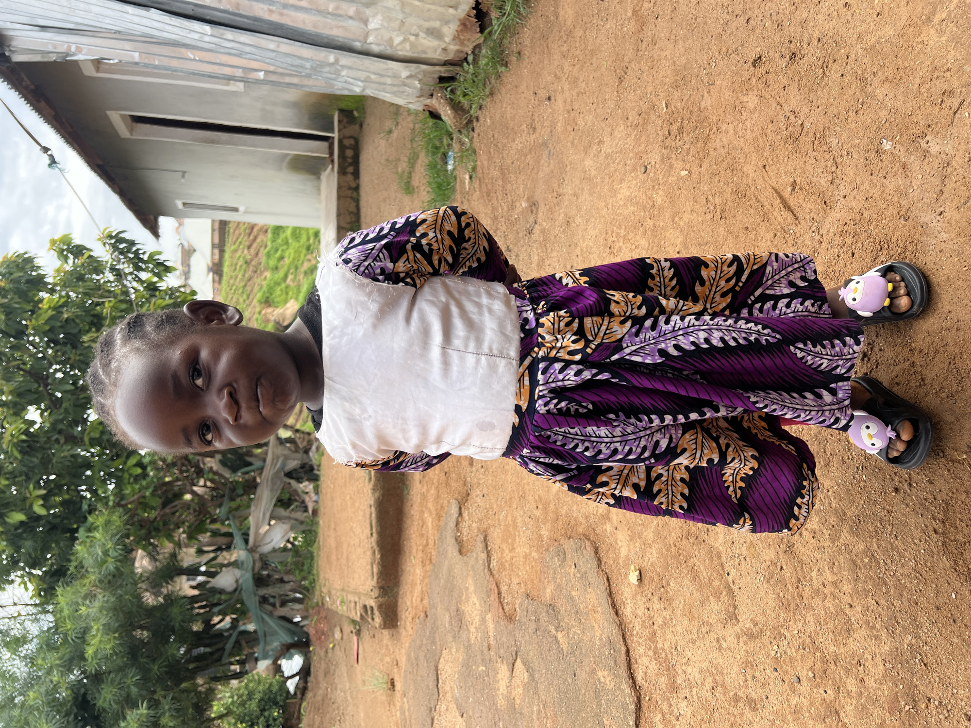 A young girl stands in an outdoor setting, likely within a residential area in Jos, Nigeria. She is facing forward, her hands clasped behind her back. The girl is wearing a white top with patterned sleeves and a purple and orange patterned skirt. She has on black sandals with light purple decorative elements on each. The ground is a reddish-brown dirt, with some scattered debris and uneven patches. In the background, there are trees, a simple building with a corrugated metal roof, and a grassy slope with what appear to be more structures in the distance. The sky is overcast.