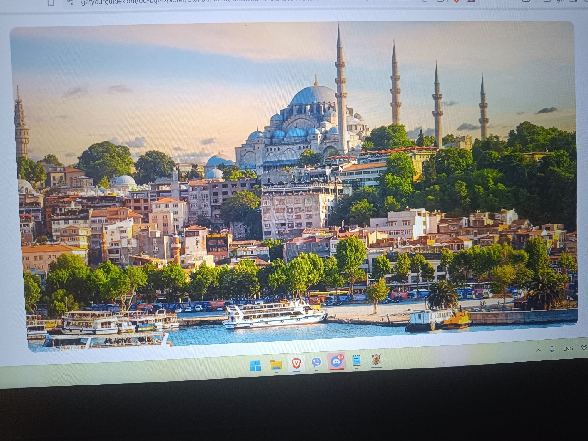 The image depicts a cityscape with a large mosque dominating the background. The mosque has a large central dome and several smaller domes, along with multiple minarets. The foreground is filled with buildings of varying heights and architectural styles, interspersed with trees. A body of water, possibly a river or bay, is in the lower part of the image with several boats docked along the shore. Cars are visible parked near the waterfront. The sky is bright with some clouds. The URL suggests the location is Istanbul. There is no evidence that this image is of Seewald, Germany.