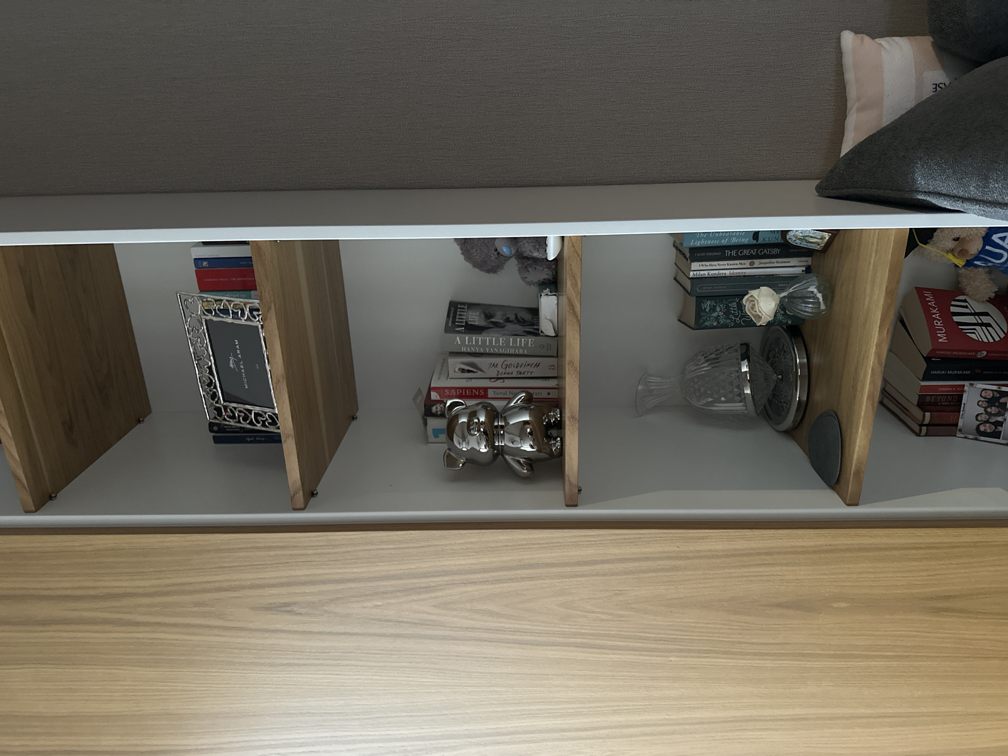 A low-angle, interior shot reveals a white shelf unit divided into three sections. Each section contains various items.
In the left section, there are several books and a silver picture frame. The middle section has more books, a teddy bear, and a silver bear figurine. The right section contains books, a flower, and a decorative silver item.
The shelves themselves are constructed with light-colored wooden supports. Below the shelf unit, there is a drawer, the front of which is made of the same light-colored wood. The background is a neutral gray.