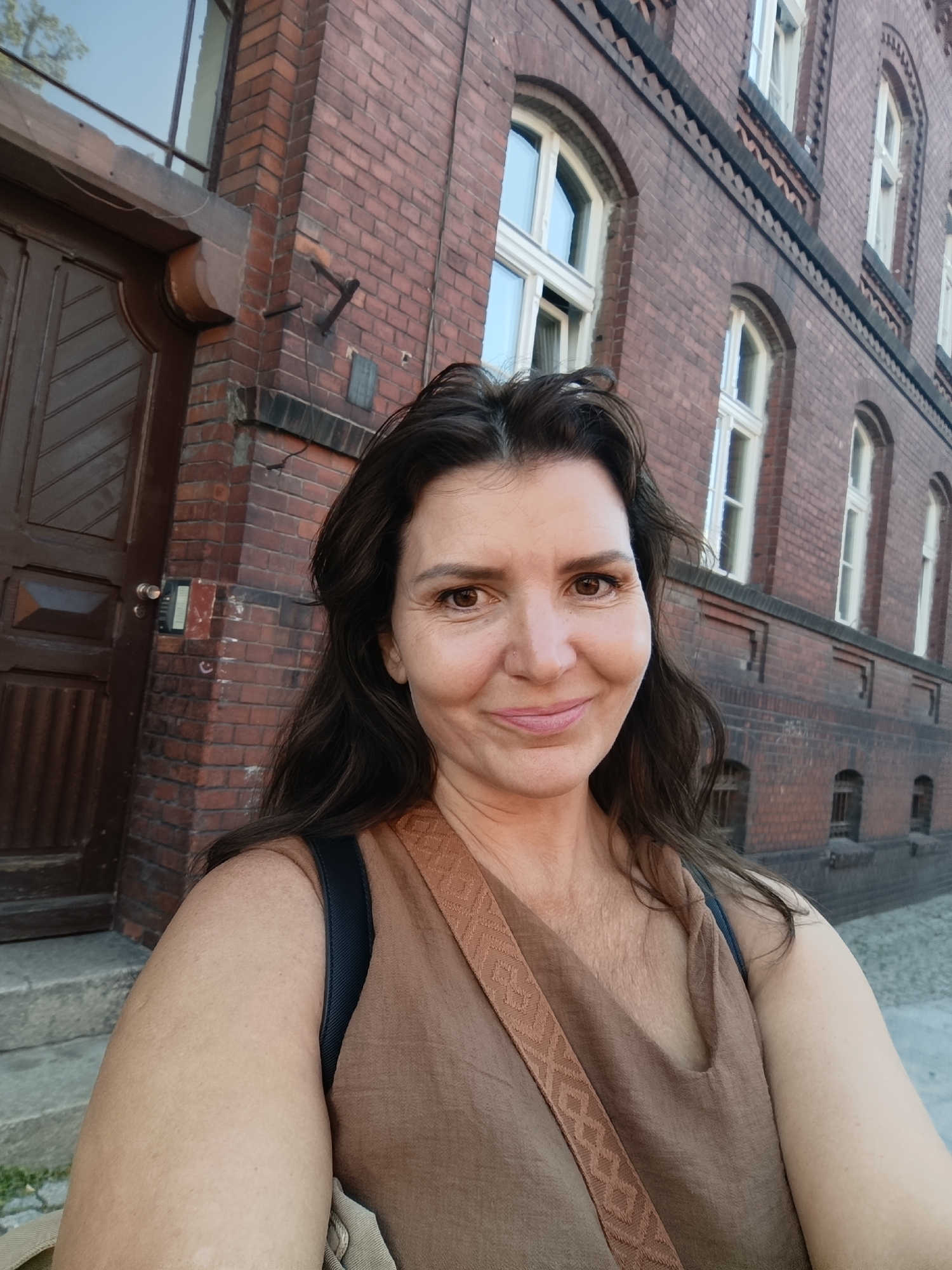 The image is a close-up, selfie-style photograph of a woman with long, dark brown hair, standing in front of a red brick building with arched windows. She is wearing a brown sleeveless top with a decorative strap. The building appears to be historic, with a large wooden door on the left side of the frame and visible steps leading up to it. The context provided suggests the location is the city of Toruń, Poland. The lighting indicates it is daytime, possibly late afternoon or early evening, given the soft illumination and the shadows. The woman is smiling gently at the camera. The background is dominated by the textured brickwork of the building and the architectural details of the windows.
