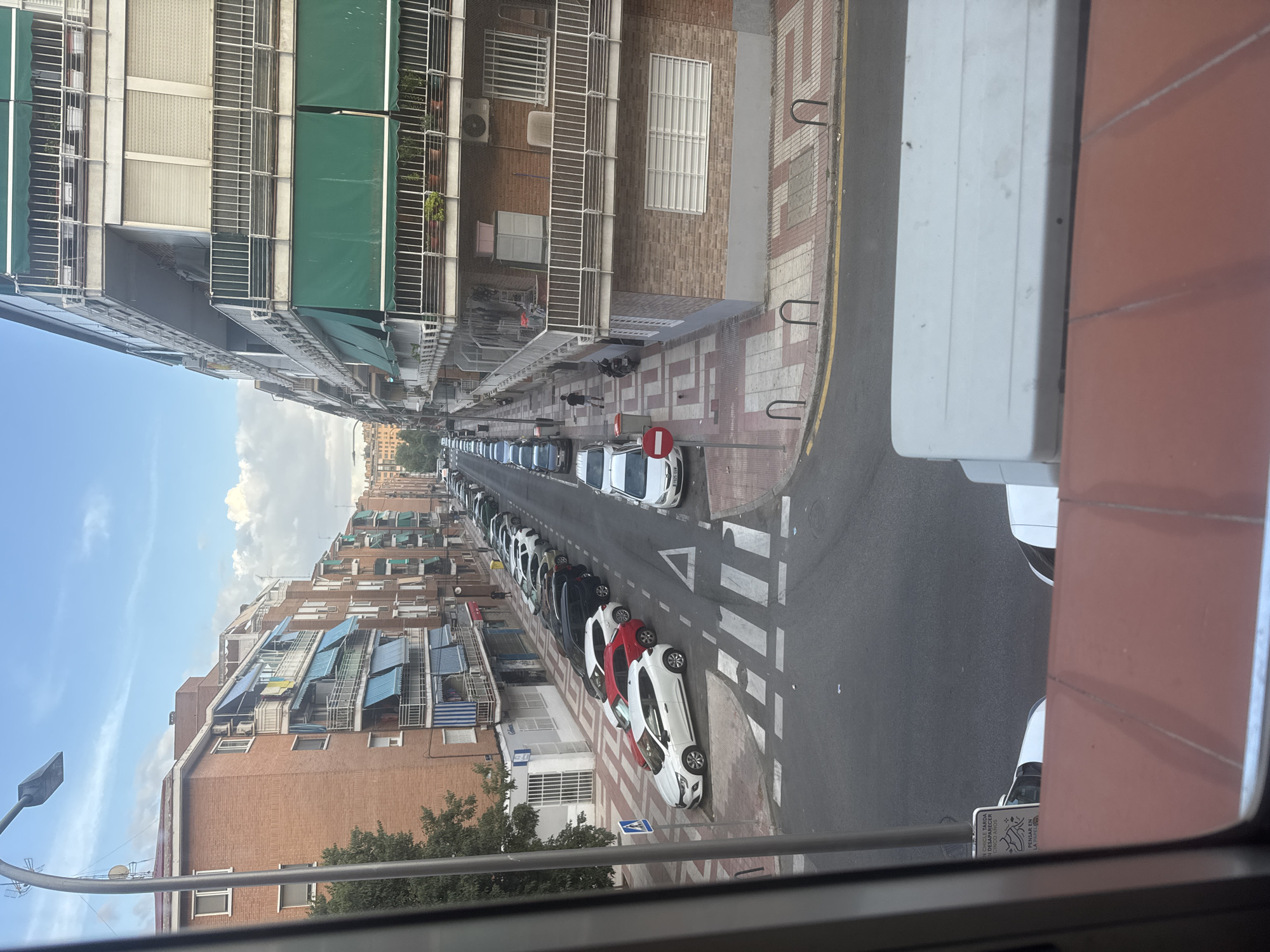 The image shows a street scene in Madrid, Spain, on a cloudy day. The perspective is from an elevated position, looking down on a street lined with multi-story brick buildings. The buildings have balconies with awnings and railings. The street is occupied by parked cars on both sides, and a few cars are moving. There is a pedestrian crossing marked on the road. Sidewalks are visible with tiled patterns. A person is walking on the sidewalk in the middle distance. The sky is blue with white clouds. The scene depicts a typical urban residential area.