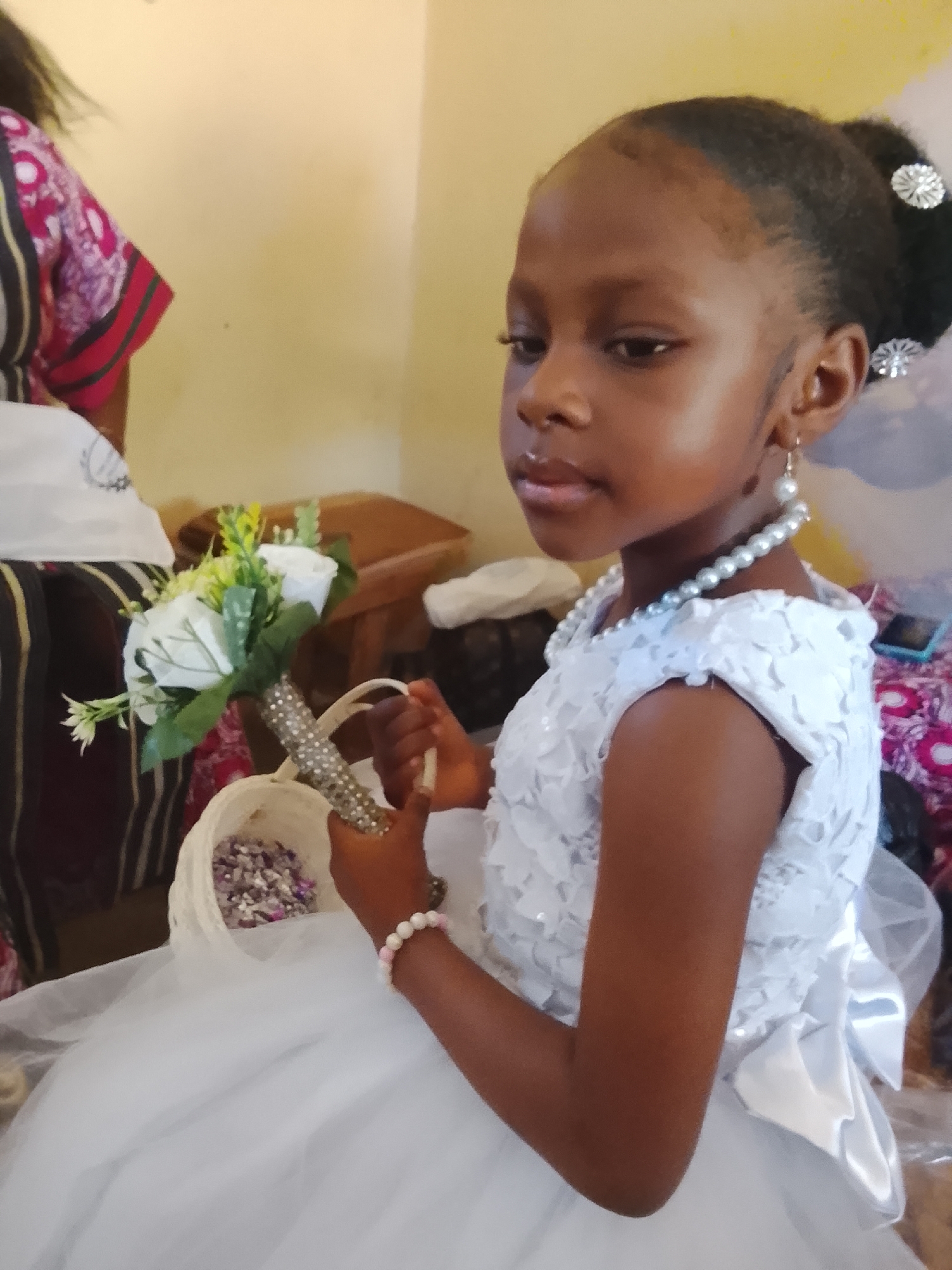 A young girl wearing a white dress, pearl necklace and pearl earrings is holding a flower arrangement and a basket in an indoor setting. Her hair is in a bun with decorative hairpins. She appears to be at a wedding, possibly as a flower girl. The city and region are unspecified.