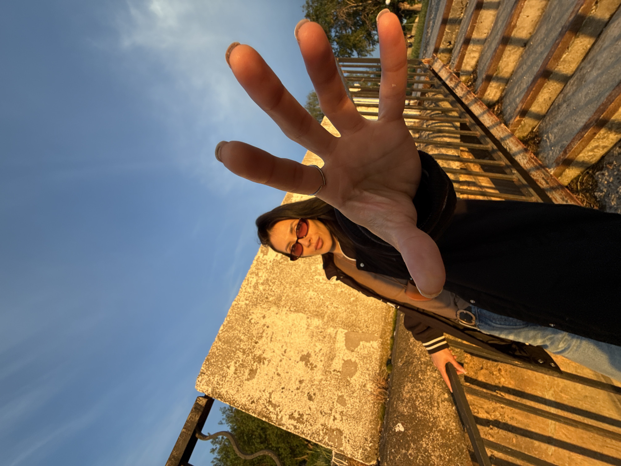 A low-angle shot depicts a woman with her hand outstretched towards the camera, fingers spread. She is wearing reddish-tinted sunglasses, a black jacket with white stripes on the sleeves, a light-colored shirt, and blue jeans. A silver ring is visible on her finger.
The background consists of a blue sky, trees, and what appears to be a concrete structure. The structure has an aging surface with visible imperfections. A metal railing runs along the edge of the structure. The sunlight is strong, creating shadows and highlighting certain textures. The location is unidentifiable.