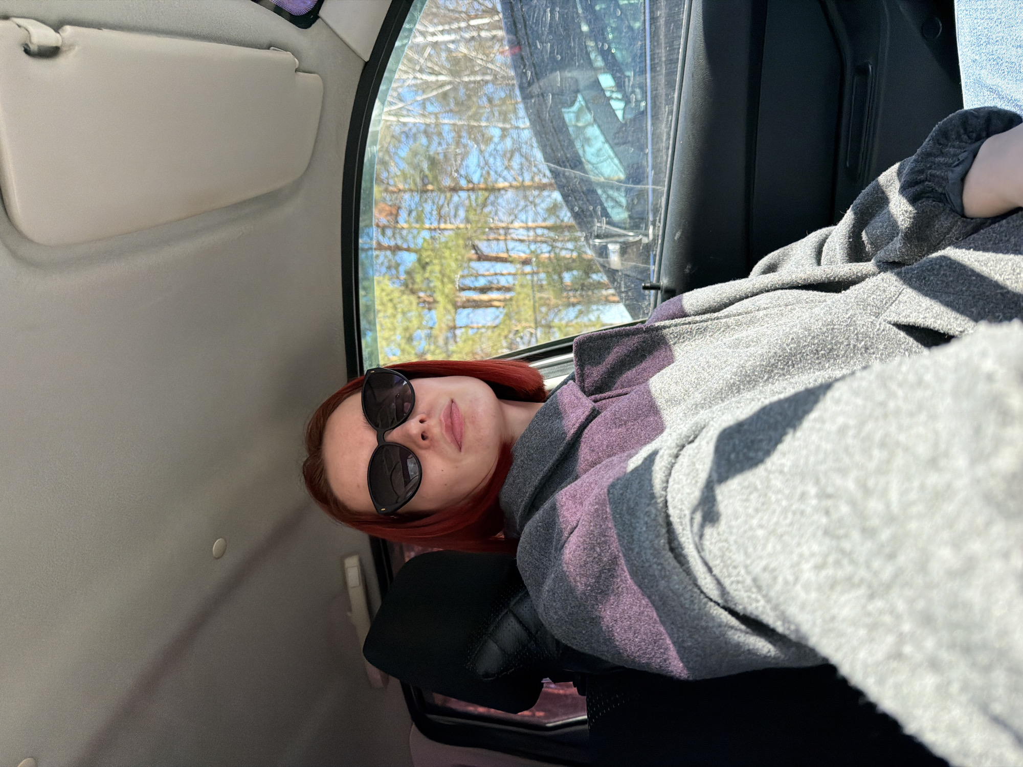 A woman with red hair and sunglasses is sitting inside a vehicle. She is wearing a gray and purple striped jacket. The vehicle is likely moving as seen from the trees and buildings through the window. The specific location in the city of Chaikovsky, Russia, cannot be determined from the image.
