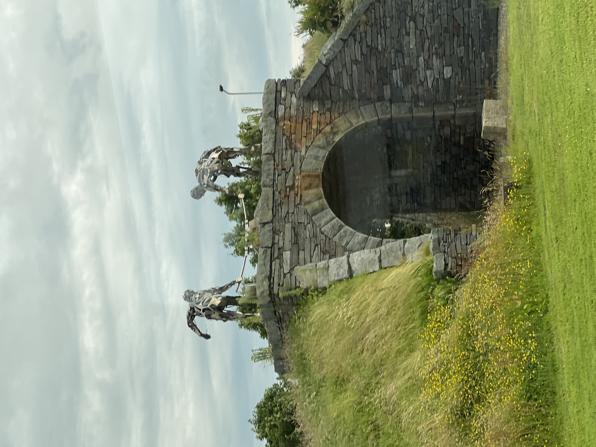 Two abstract sculptures of human figures, appearing to be made of welded metal, stand atop a stone archway bridge. The figures are positioned on either side of the apex of the bridge, facing each other and holding long poles or staffs. The sculpture on the left is in a more upright stance, while the one on the right is bent forward. The bridge is constructed of rough-hewn, grey stones with some reddish-brown stones incorporated into the arch. The area surrounding the bridge is covered in green grass, with a slope covered in tall, wispy grass and scattered yellow wildflowers on the left side. A paved pathway is visible beneath the arch. In the background, trees and foliage are present, and the sky is overcast with grey clouds. A single lamppost is visible on the right side of the bridge. This scene is located at the entrance to Letterkenny Rugby Club in Letterkenny, County Donegal, Ireland.