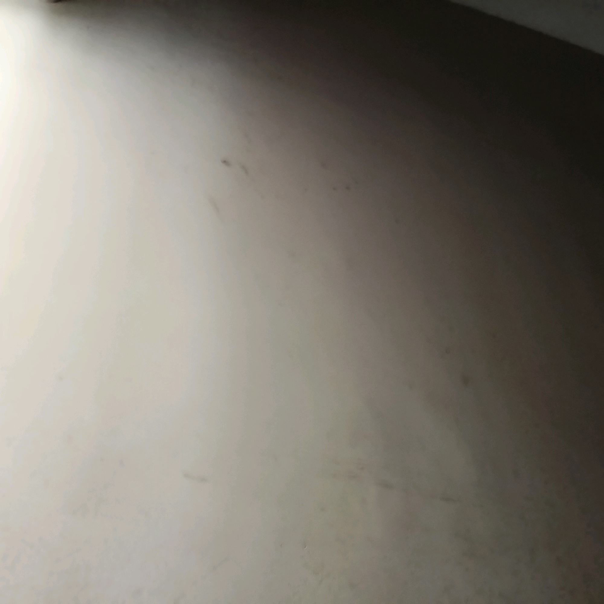 The image displays a close-up, angled view of a textured surface, likely a wall or floor, exhibiting a gradient of light and shadow. The dominant color is a light, neutral tone that transitions to a darker shade in the upper right corner, suggesting the presence of an overhead light source or a shadowed area. Subtle variations in texture and scattered, small dark marks are visible across the surface, hinting at minor imperfections or dust. The scene is devoid of any identifiable objects, people, or specific environmental features that would indicate a precise location within Bengaluru, India.