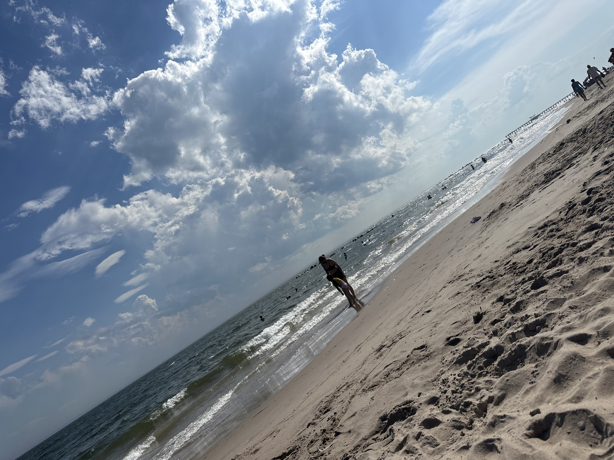 The image depicts a beach scene on a sunny day with a wide expanse of sand and the ocean. The sky is partly cloudy, with large cumulus clouds and some wispy cirrus clouds. The ocean is a dark blue-green with gentle waves breaking on the shore. Several people are visible on the beach and in the water, engaging in activities such as walking, swimming, and potentially playing. In the foreground, a person is walking along the wet sand near the water's edge, casting a shadow. In the middle ground, more figures are scattered along the beach and in the shallow water. A long pier or boardwalk extends into the ocean in the distance, with more people visible on it. The sand is a light brown color and appears to be fine-grained. The overall impression is of a typical summer day at a popular beach. The angle of the shot is tilted, giving a dynamic perspective of the scene. This scene is consistent with the beaches found in Queens, New York, such as Rockaway Beach.