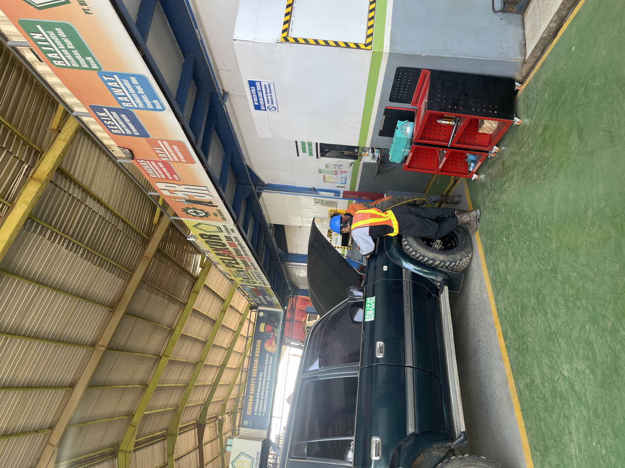 An outdoor, covered automotive service bay in Unknown, Indonesia features a dark green four-wheel-drive vehicle with its hood open. Two individuals, one wearing a blue hard hat and a high-visibility orange and yellow vest over a light-colored shirt and dark pants, and another person in dark clothing, are attending to the vehicle. The bay has a corrugated metal roof supported by yellow metal beams. A large banner with Indonesian text about safety and workplace standards hangs along the upper wall. Signs with text like "RAJIN," "BERSIH," and "RAWAT" are visible. The floor is painted green with a yellow border. In the background, a red truck is partially visible. To the right, a white wall has signage for "WORKSHOP ADMINISTRASI" and a tool cart with red and black shelves holding various items, including blue and green containers.