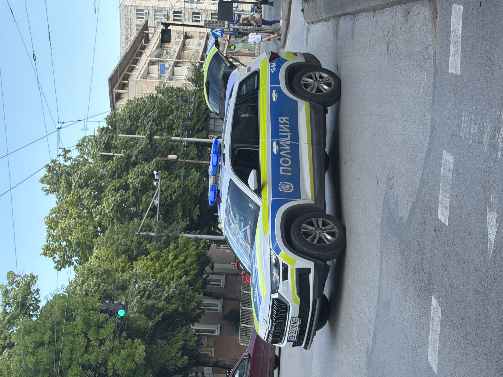 A daytime, street-level shot depicts a white and blue police vehicle with "ПОЛИЦИЯ" (Police) printed in Cyrillic on its side. The vehicle's rear hatch is open, and blue emergency lights are visible on the roof. The license plate, EH2202ME, is visible on the front of the car. The car is parked on a grey asphalt road with white lane markings. Trees and buildings are visible in the background, and a green traffic light is noticeable hanging above the street. Pedestrians are walking on the sidewalk in the distance. The scene is in Pleven, Bulgaria.