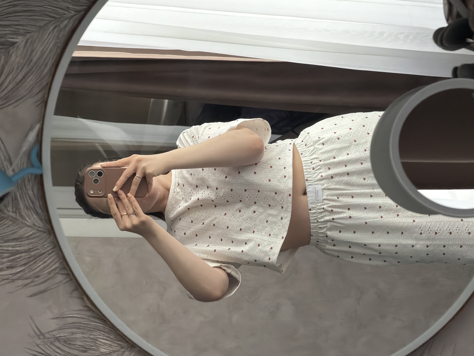 A woman is taking a selfie in a mirror. She is wearing a white pajama set with red hearts. The mirror is round and has a decorative border. A phone and ring light are visible. A window is visible in the background. The location is Reshetnikovo, Russia.