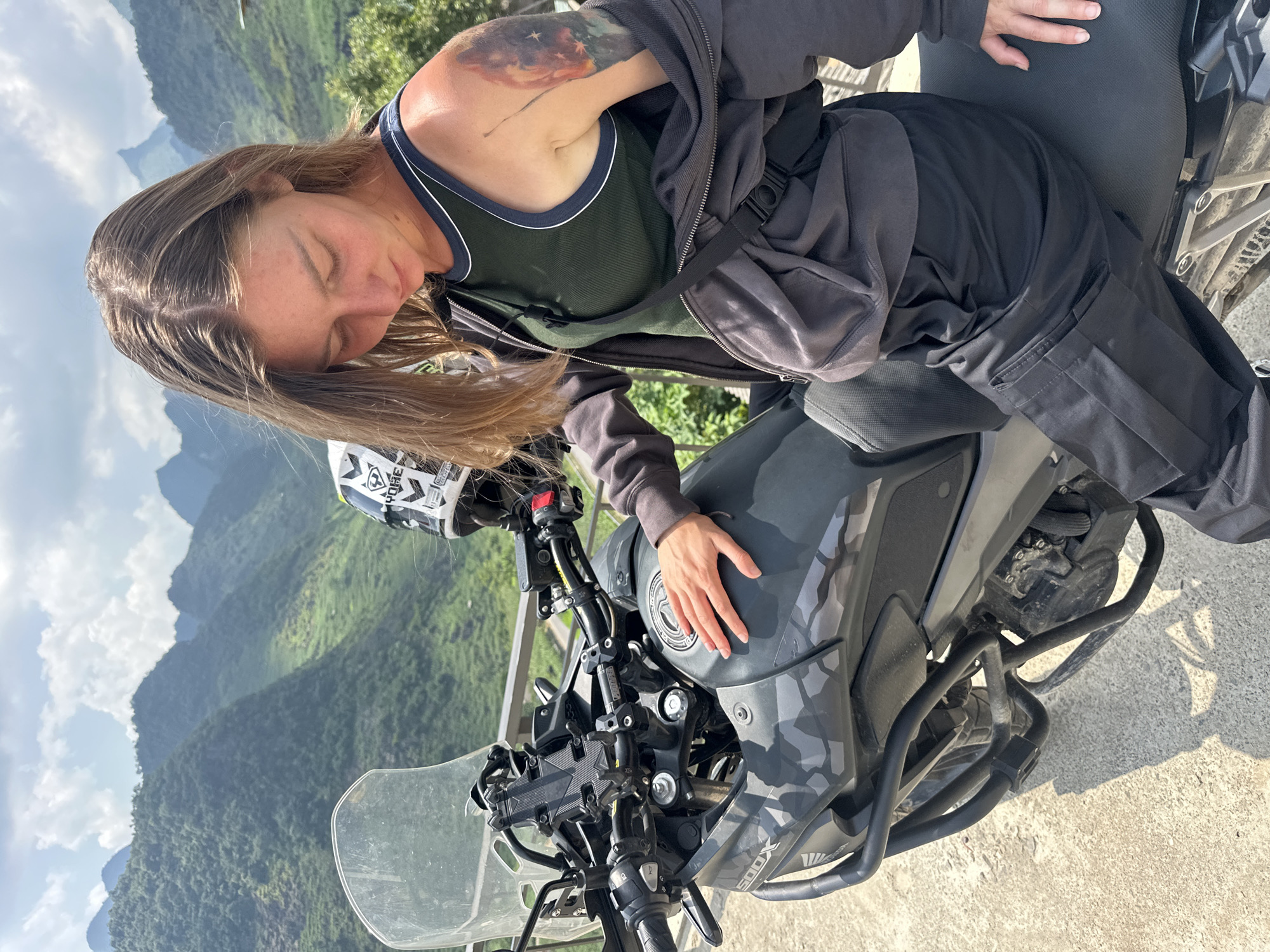A woman with long brown hair and a tattoo on her left arm is resting on a gray and black camouflage motorcycle in Lũng Phìn Commune, Vietnam. She wears a dark green tank top with white trim, a dark gray hoodie, and dark pants. Her eyes are closed, and she rests her head on the fuel tank with her right hand on top. The motorcycle has a helmet resting on the left handlebar. A clear windscreen is visible. In the background, a mountain range extends into the distance. The sky is partly cloudy.