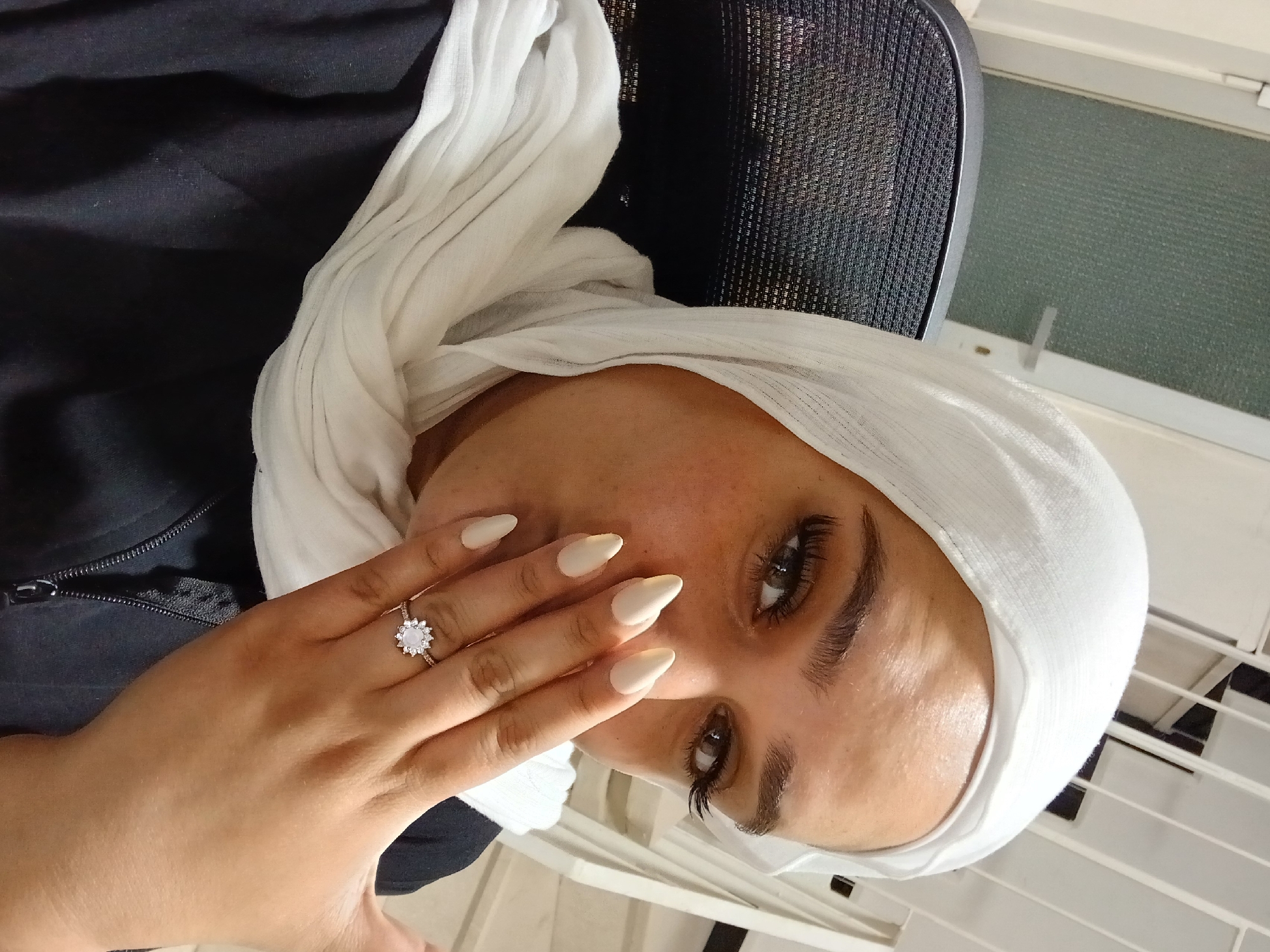 A medium shot shows a woman wearing a white hijab and a black jacket. She is seated in an office chair, with her hand covering her mouth. She is wearing a ring with a large, clear stone surrounded by smaller stones on her ring finger. Her nails are painted a pale cream color. The background appears to be an indoor setting, possibly an office, with a door visible in the upper left corner. The lighting suggests it is daytime, possibly artificial indoor lighting. There are no discernible features to place this image in Rukn ad-Din Municipality, Syria.