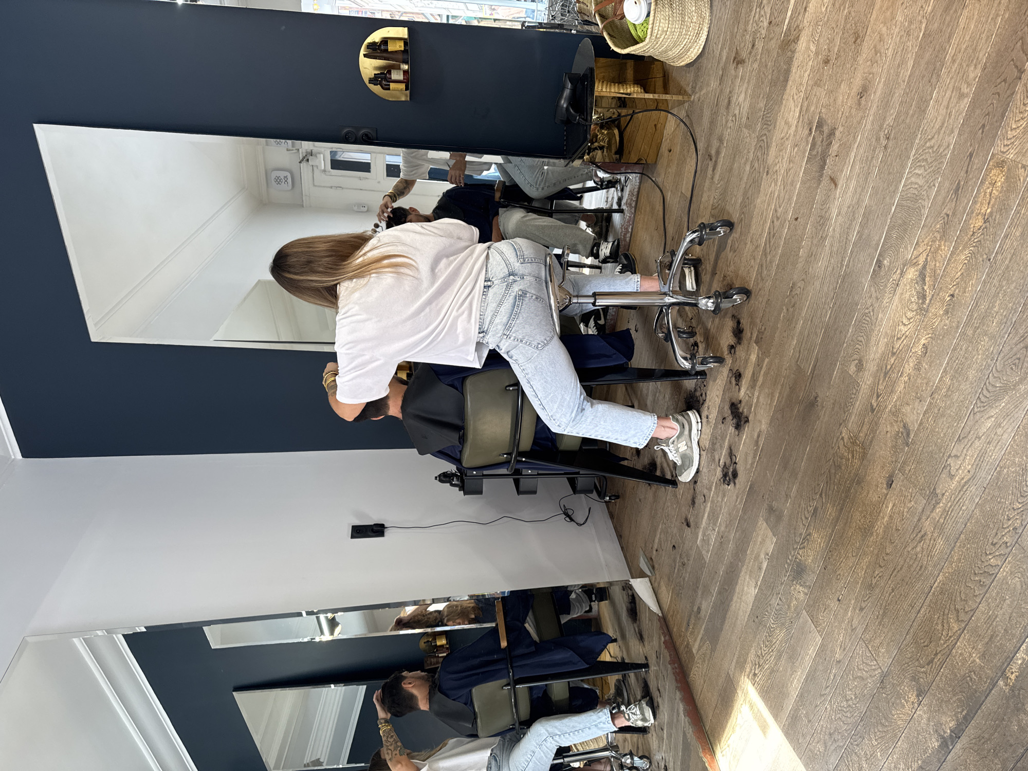 In a well-lit, modern barbershop in Lille, France, a female hairdresser with blonde hair is seated on a rolling stool, facing away from the camera. She is wearing a white t-shirt and light-wash jeans. Her hands are raised, and she appears to be actively cutting the hair of a male client who is seated in a chair in front of her, also facing away. The client is wearing a dark blue cape. The floor is made of distressed wooden planks and is visibly scattered with dark hair clippings, indicating a recent haircut.
To the left of the main scene, another mirror reflects a similar setup, with another hairdresser attending to a client. Multiple large mirrors line the walls, creating a sense of depth and reflecting the interior of the establishment. A black electrical outlet with a cord is visible on the navy blue wall.
To the right, a gold-colored shelf is mounted on the navy blue wall, holding small amber bottles. Below this shelf, a small, round gold table holds a black hairdryer. A woven basket containing what appears to be a coffee cup and other items sits on the floor next to the table. The overall environment suggests a busy, functional hair salon.