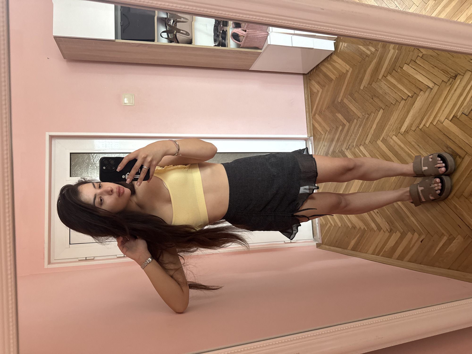 A young woman is captured in a mirror selfie, posing indoors in what appears to be Plovdiv, Bulgaria. She is seen holding a black smartphone with a prominent camera array and a case featuring some white text or logo, likely taking the photo. Her long, dark hair cascades over her shoulders and arm. She is wearing a yellow ribbed crop top, a black ruffled mini-skirt, and brown platform sandals, with white painted nails visible on her toes. A silver-toned watch is on her left wrist, and she has a ring on her right hand.
The setting is a room with light pink walls. A light-colored, possibly wooden, shelf unit with white compartments is mounted on the wall behind her reflection, holding various items like bags, one of which is a light pink woven bag. A light switch or outlet is also visible on the pink wall. The floor has a distinctive light brown wooden herringbone pattern. A white door or window frame is partially visible behind the woman. The overall lighting suggests a bright, possibly daytime, interior. The image is presented as a horizontal mirror reflection, suggesting the original capture was oriented this way or has been rotated.