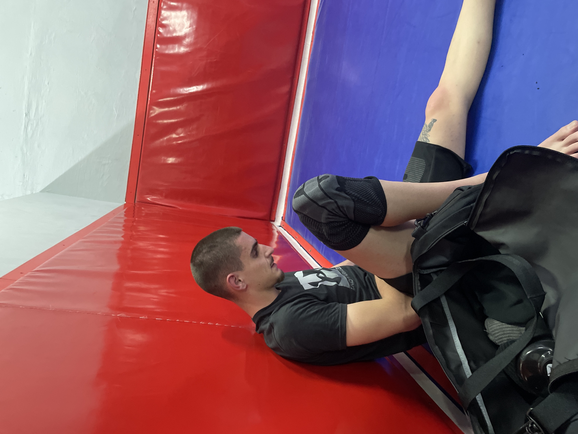 The image shows a man lying on a red and blue padded mat. The mat is in front of a wall. The man is lying on his side, facing left. He has a shaved head, a light mustache, and a dark t-shirt. He is wearing a knee brace on one knee. A black gym bag sits next to his legs. The environment is a gym. It is not possible to tell the specific location in Plovdiv, Bulgaria from the image.