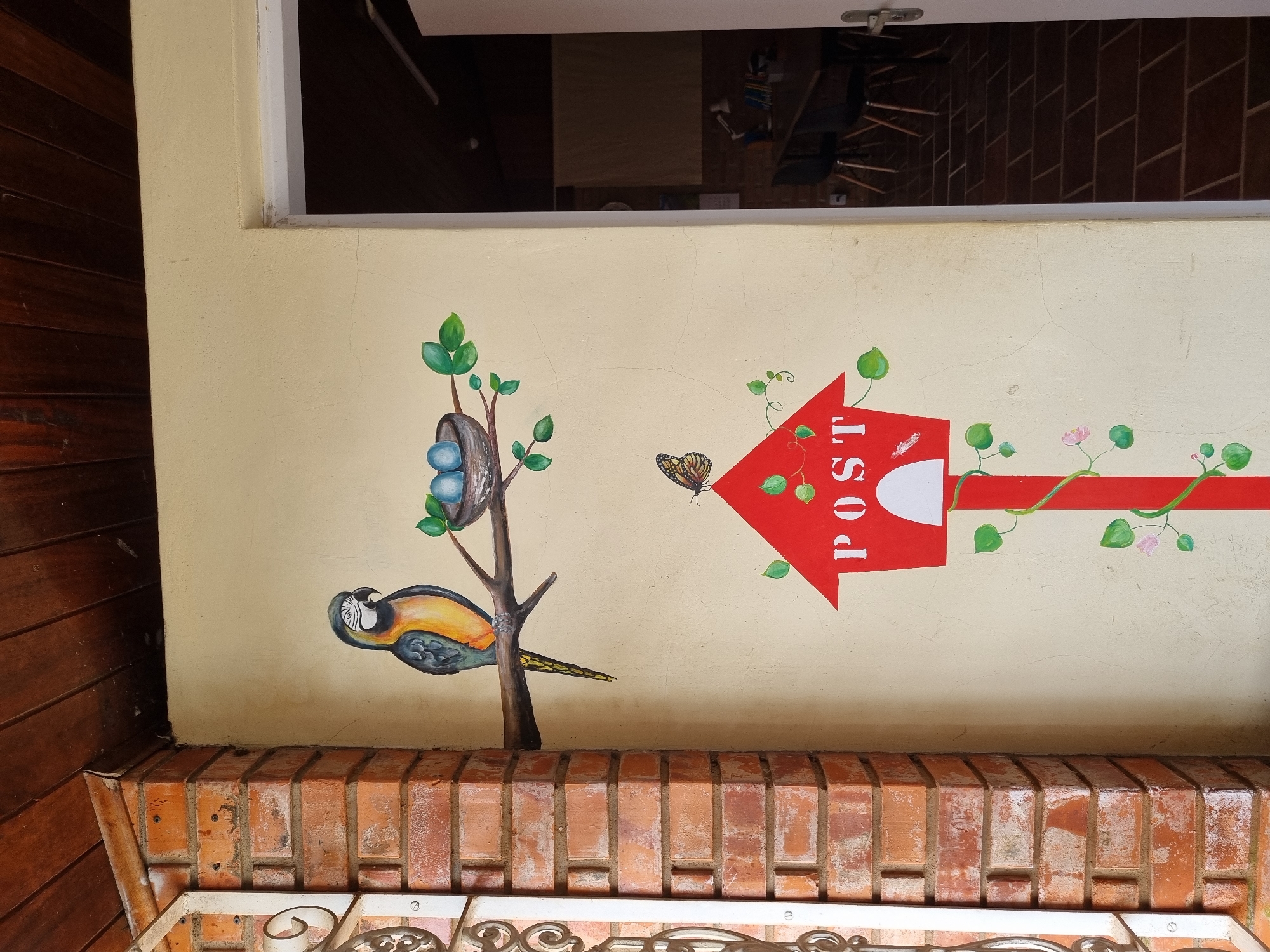 The image shows a mural painted on a light-yellow, textured exterior wall. To the left, a brick pillar with weathered red bricks is visible, along with a white metal decorative railing. The mural features a colorful parrot with blue, yellow, and green plumage perched on a brown branch. Next to the parrot is a bird's nest containing three light-blue eggs. Below the branch, a red mailbox is painted in the shape of a house with a white opening. The word "POST" is stenciled in white letters above the opening. A monarch butterfly with orange and black wings is painted above the mailbox. Green vines with leaves and small pink flowers twine around the mailbox and its red post. The ceiling above is made of dark wooden planks. To the right of the wall, a doorway leads into a room with dark tile flooring and a glimpse of a table and chairs. The scene appears to be an exterior wall of a building, possibly in a residential or commercial area in Laranjal Paulista, Brazil, judging by the architectural style and the commonality of such artistic embellishments in Brazilian cities. There are no people or animals present in the image, only the painted elements. The lighting suggests it is daytime, with natural light illuminating the scene.