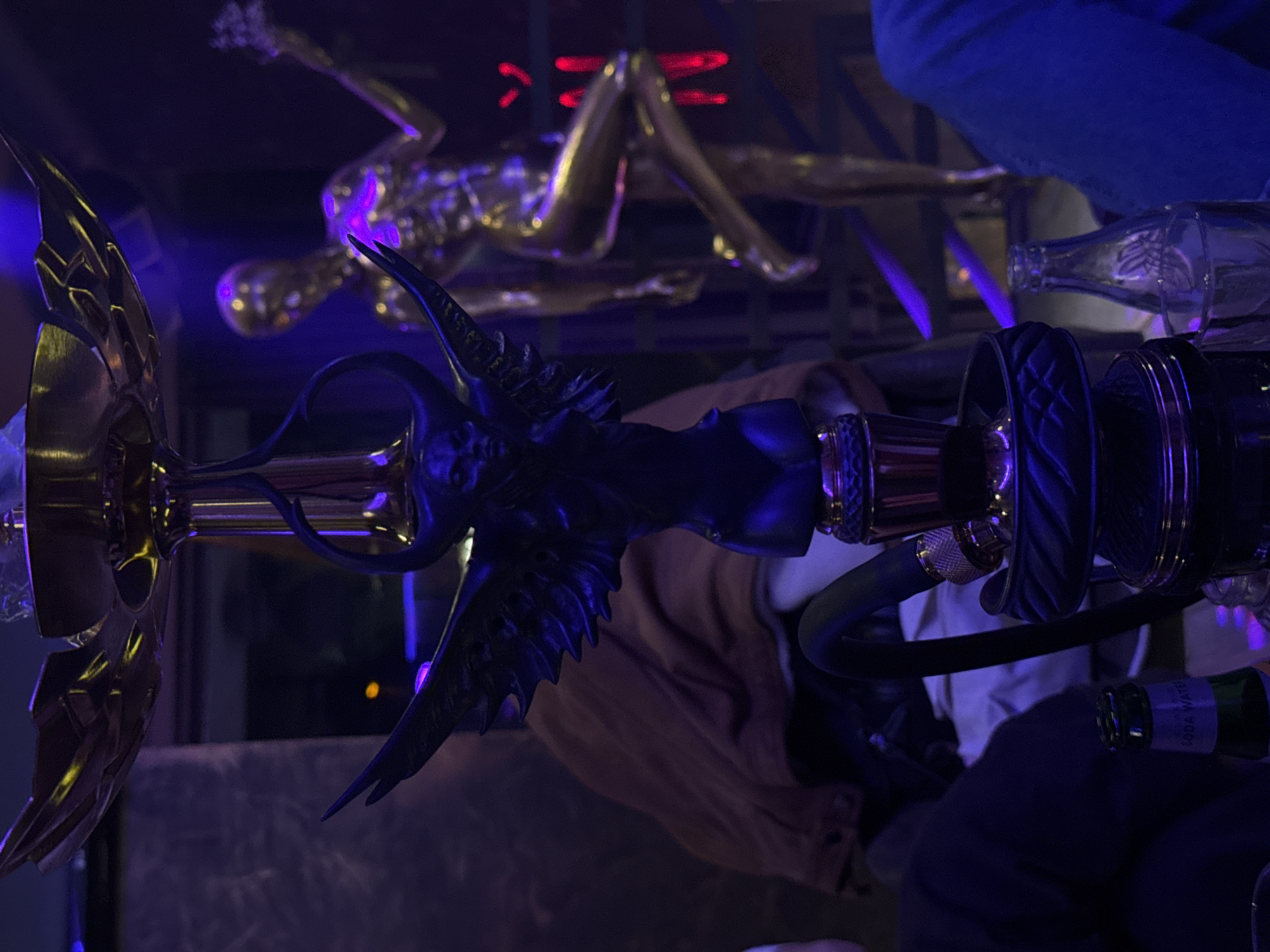 The image depicts a hookah with a gold and black design. A golden statue of a reclining nude woman is in the background, illuminated with purple light. The setting appears to be a dimly lit indoor space. A glass bottle and a person's legs in blue jeans are also visible. There is not enough information to determine if the image was taken in the city of Burgas, Bulgaria.