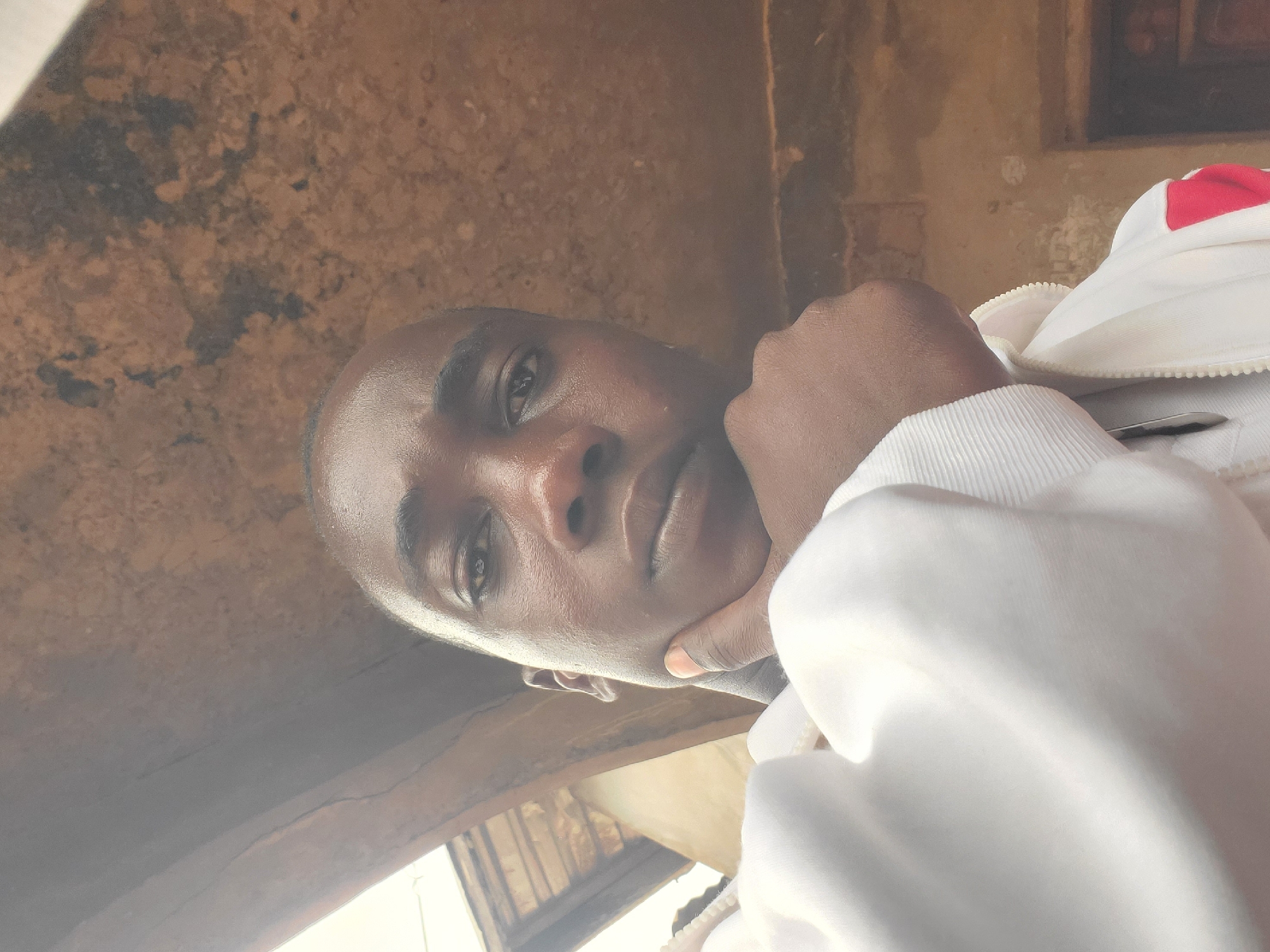 This is a selfie of a young Black male with short hair and dark skin. He is resting his chin on his hand, with his elbow on his chest. He is wearing a white hooded jacket with red and white detailing on the sleeves and zipper. The background appears to be an outdoor setting with weathered, brown, textured walls and a wooden roof structure overhead, consistent with the architecture found in Nsugbe, Nigeria. The lighting suggests it is daytime, with some light filtering in from the side.