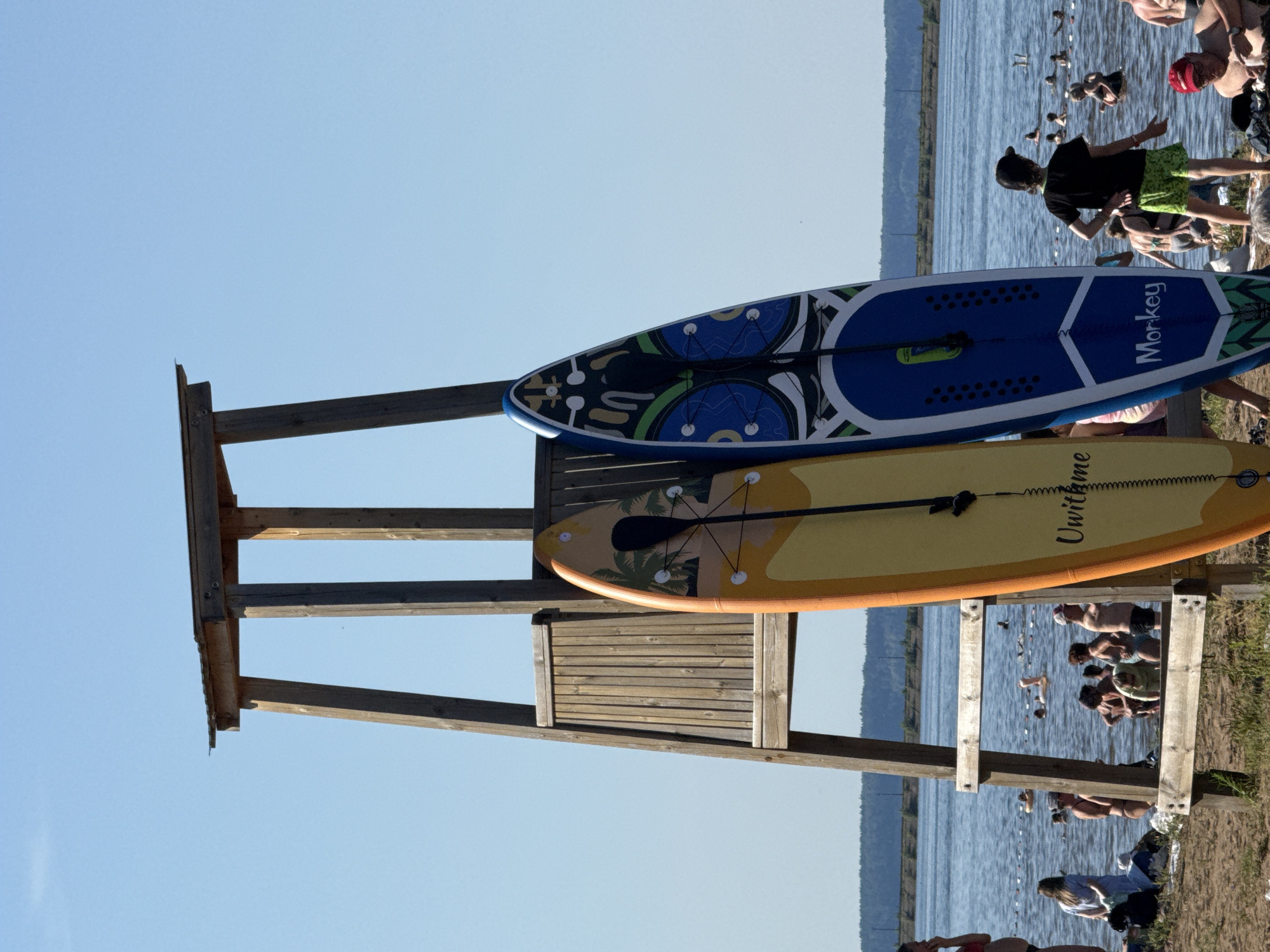 The image depicts a sunny day at a beach in Chaikovsky, Russia, with a prominent wooden lifeguard tower in the background. Two stand-up paddleboards are leaning against the tower, one orange with a palm tree design and the other blue with a geometric pattern and the word "Monkey" visible. In the water, numerous people are swimming and wading, some with inflatable rings. The beach area is populated with individuals sunbathing and standing near the water's edge. The background features a distant treeline and a clear blue sky.
