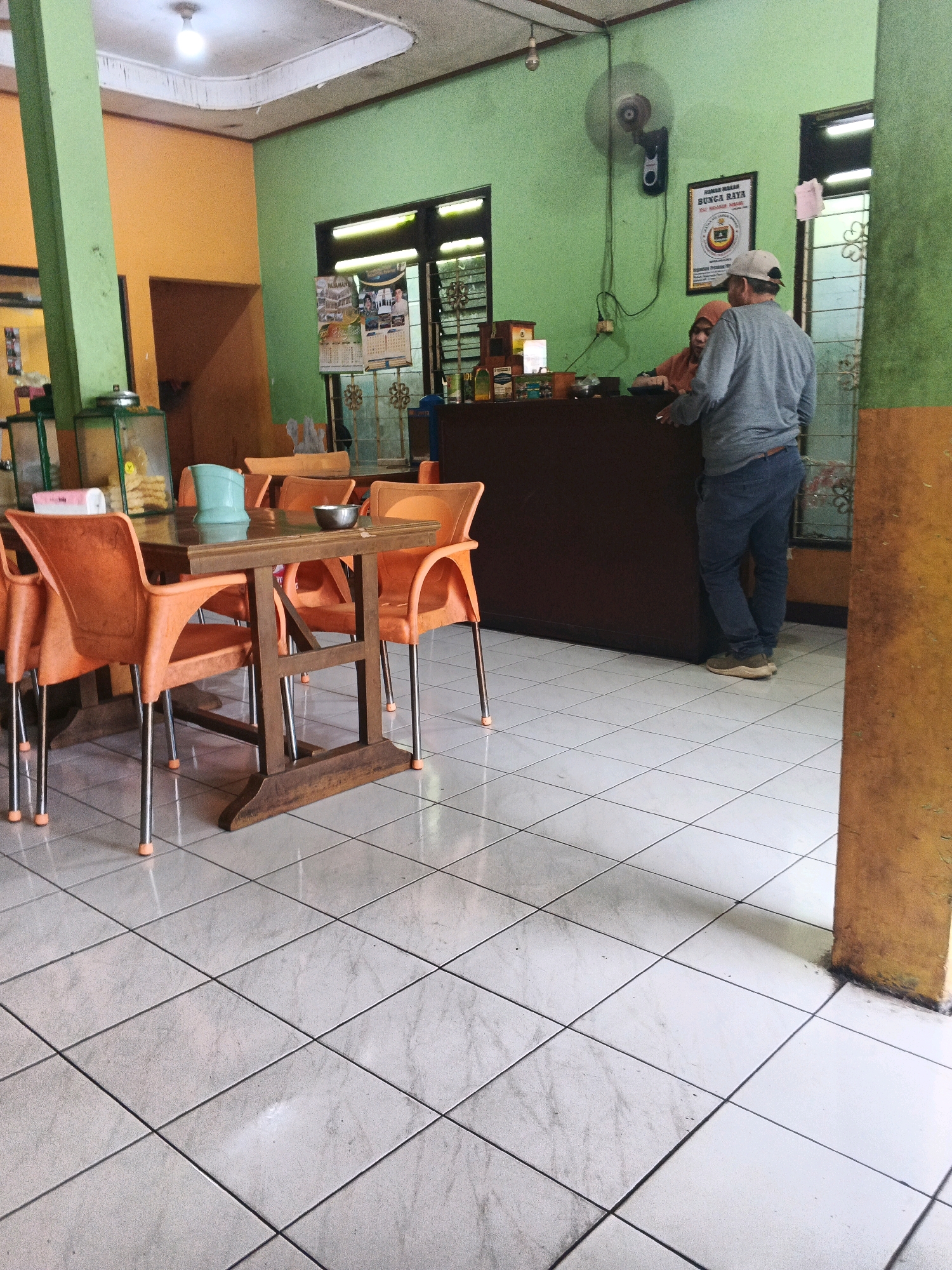 This image depicts the interior of a food stall or small restaurant in Ungaran, Indonesia. The walls are painted in contrasting bright colors: a pale orange on the left and a vibrant green on the right. The floor is tiled with large, square, pale gray tiles laid diagonally.
In the foreground, there is a wooden table with a glass top, surrounded by four bright orange plastic chairs with metal legs. On the table, a teal plastic pitcher and a small metal bowl are visible.
In the background, a dark brown counter extends across the scene. Behind the counter, two individuals are interacting. A person wearing a grey long-sleeved shirt, blue jeans, and a baseball cap is standing and facing away from the camera, looking towards the counter. Partially obscured by this person, another individual wearing an orange headscarf and a brown top is visible behind the counter, seemingly attending to the customer.
To the left of the counter, there is a glass display case with some items inside, and further to the left, a tall, bright green pillar supports the ceiling. On the green wall, a framed poster with text and images, likely advertising the establishment, is hanging. A wall-mounted fan is also visible on the green wall above the poster.
The overall environment appears casual and functional, typical of a local eatery.