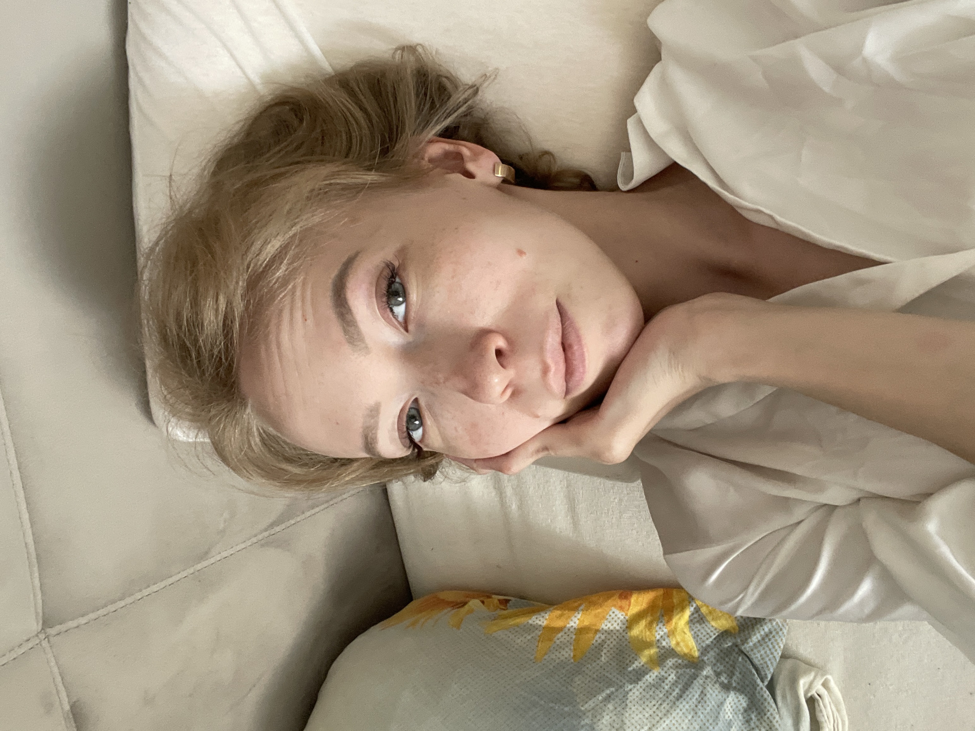 A woman with fair skin, blond hair, and visible freckles is lying on a beige sofa, propping her head with her right hand. She is wearing a white top or gown. There's a pillow with a sunflower design next to her. The location is not specified in the image.