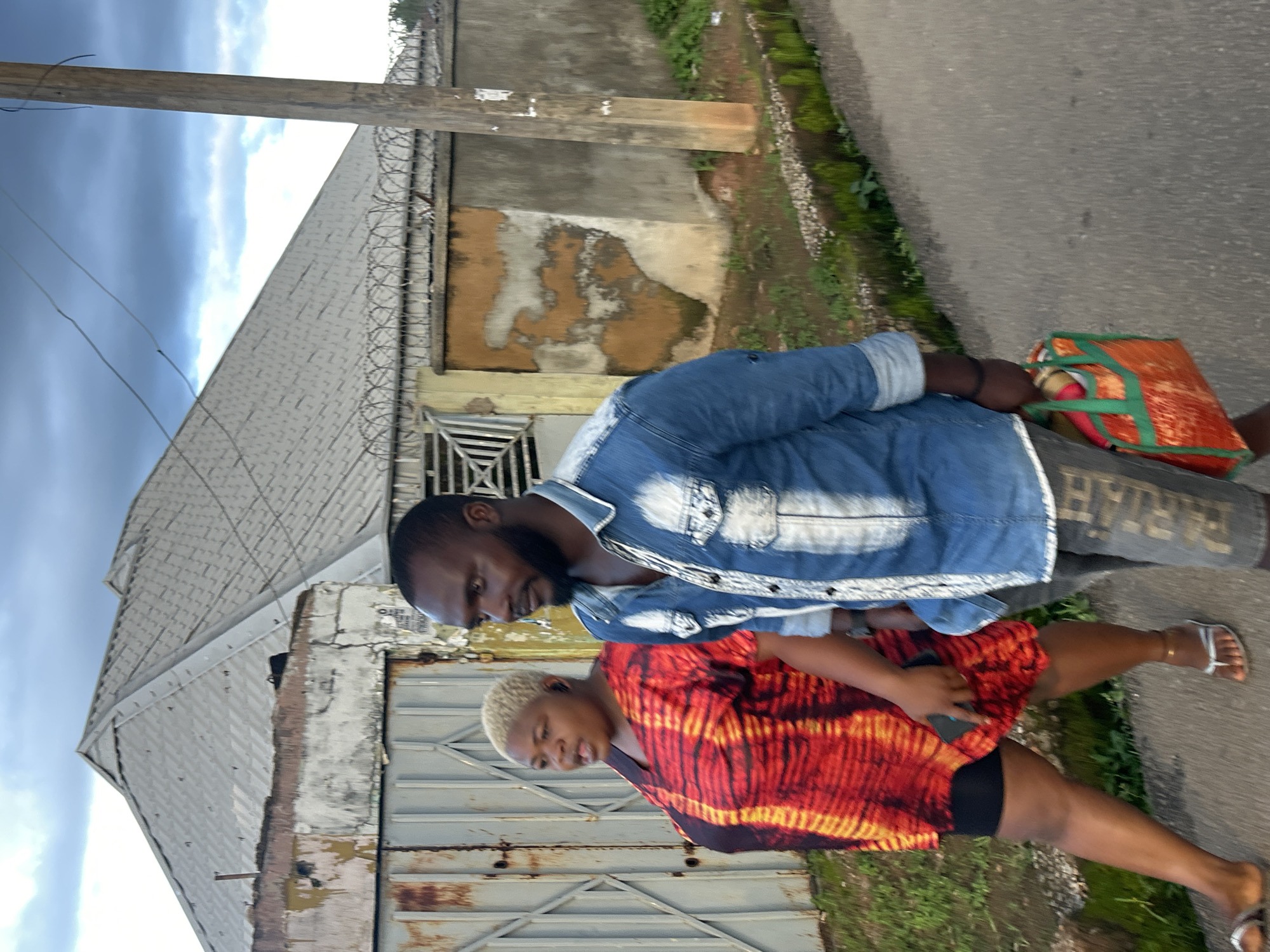 In Jos, Nigeria, a man and a woman walk along a paved road. The man, with a beard, wears a blue denim shirt and grey pants. He carries a red and green patterned bag in his right hand. The woman, with short blonde hair, wears a vibrant red and orange tie-dye dress and black shorts. She holds a smartphone in her left hand. Behind them, a building with a corrugated metal roof and a weathered, grey-toned wall is visible. Barbed wire runs along the top of the wall. A wooden utility pole stands near the road. The sky above is partly cloudy.