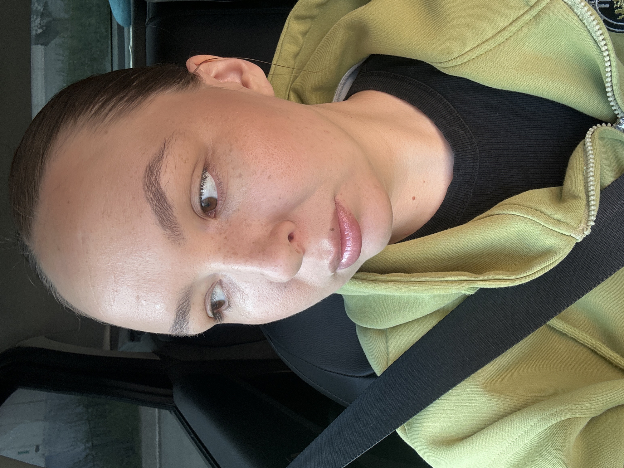 A person with light brown hair pulled back is sitting in the driver's seat of a car, wearing a light green hooded sweatshirt and a black shirt. The person is looking to the left, with their right side of their face visible to the camera. A black seatbelt is fastened across their chest. The interior of the car is dark, with a window visible on the left. The exterior seen through the window appears to be a street in Chaikovsky, Russia, with some vegetation and a building. The lighting suggests it is daytime.