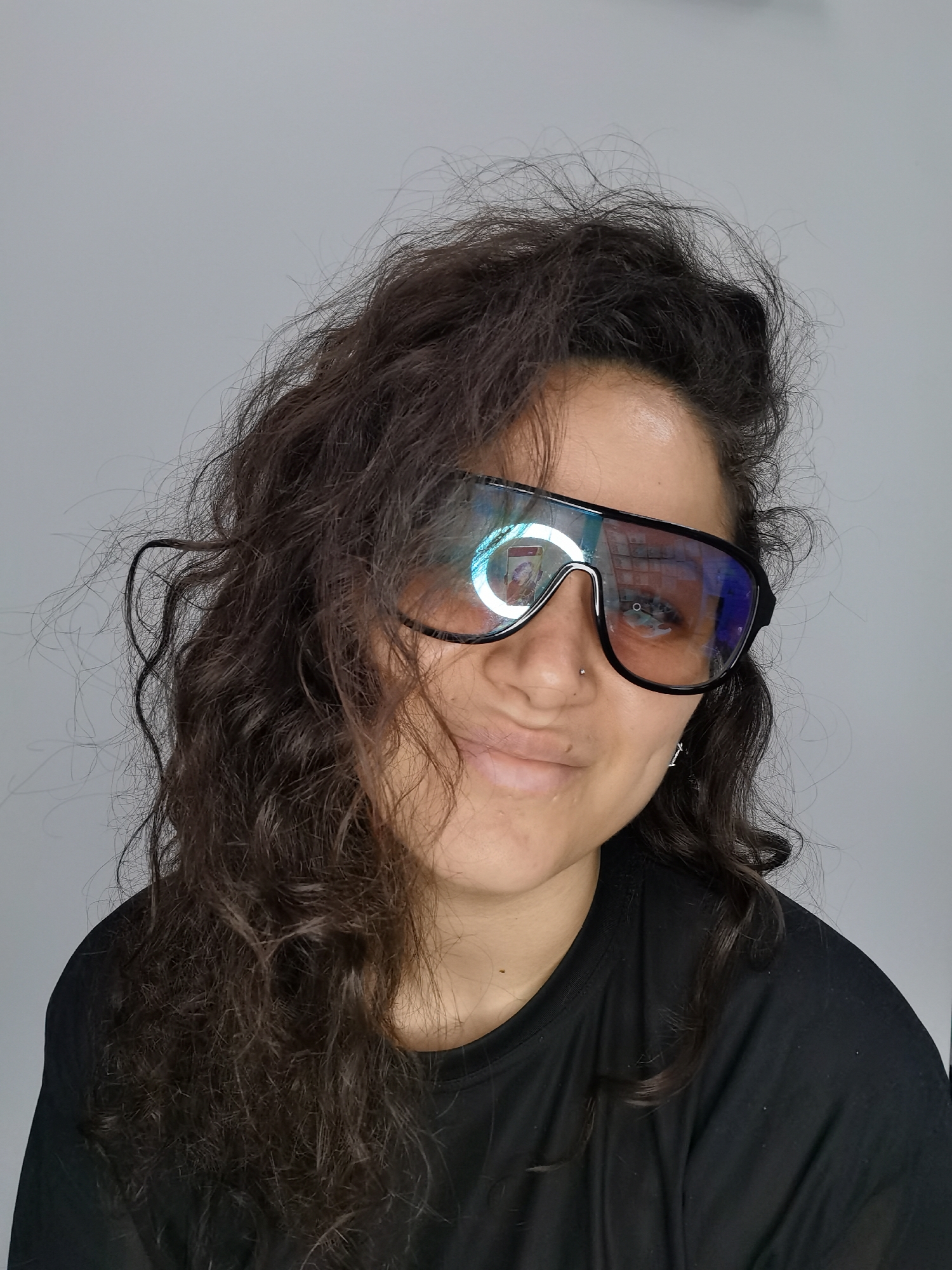 A close-up, medium shot captures a person from the shoulders up against a plain gray background, suggestive of an indoor setting. The individual, positioned slightly off-center to the right, has dark, curly hair that appears somewhat tousled. They are wearing a black t-shirt and large, black-rimmed glasses with tinted lenses that reflect a ring light and other objects. A nose ring is visible on the left side of their nose, and a small earring is on their left earlobe. The person's head is tilted slightly to their left, and they are looking towards the camera with a subtle smile. The lighting appears to be coming from the front, creating highlights on the glasses and illuminating the face. There is no indication of the city of Chaikovsky, Russia, or any specific urban or environmental context within the image.