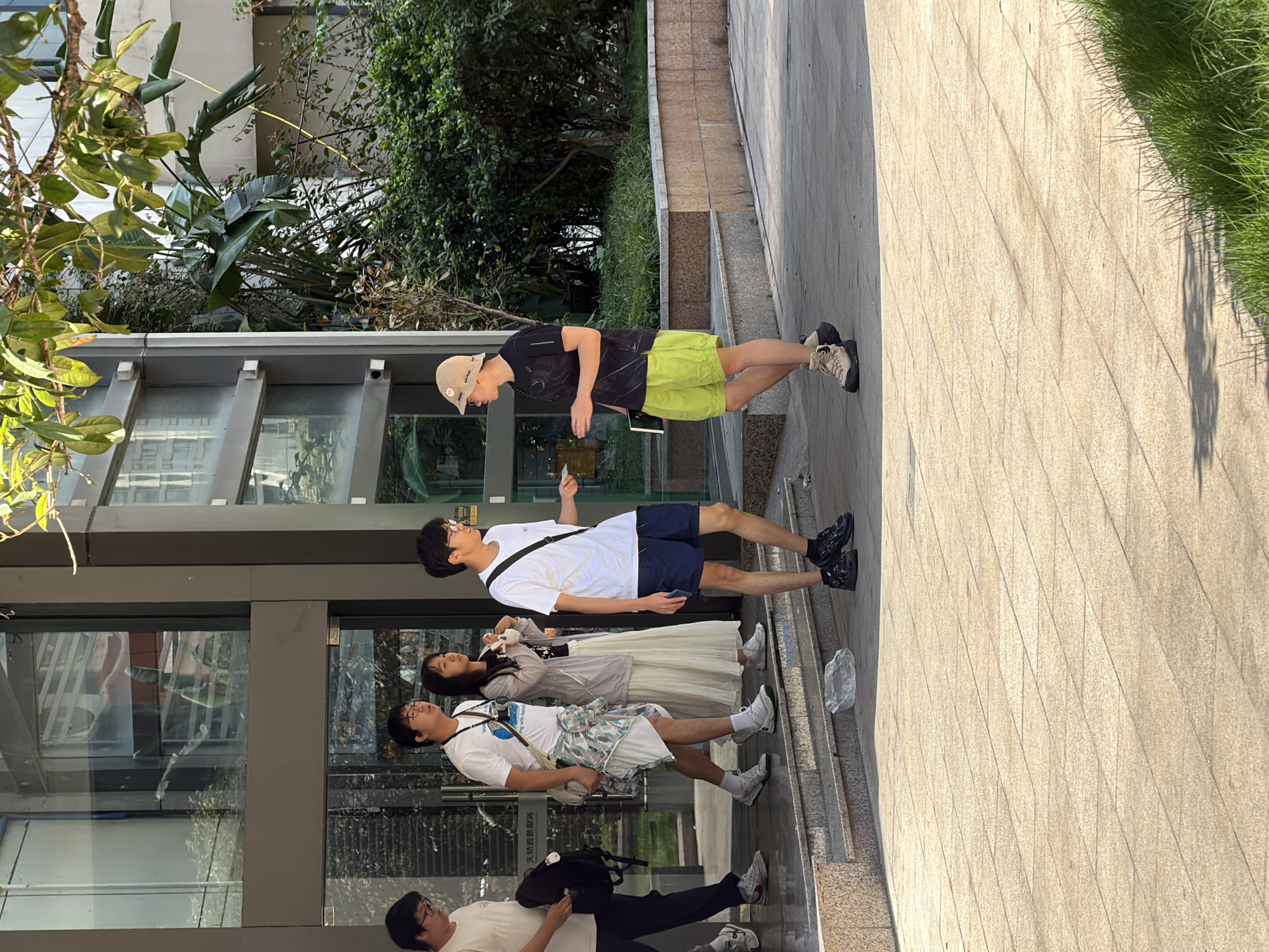 The image depicts an outdoor scene in front of a modern building with glass doors, possibly in Yuzhong District, China, given the presence of East Asian individuals. Sunlight casts distinct shadows on the tiled pavement.
In the foreground, two young men are interacting. One, wearing a black t-shirt with a patterned design and bright neon yellow shorts, stands with his left hand outstretched, holding a small object, possibly a piece of paper. He is also wearing a tan bucket hat and beige hiking boots. The other man, in a white t-shirt and dark blue shorts, stands facing him, looking down at the object in the first man's hand. He is wearing black athletic shoes.
Behind them, on a set of concrete steps leading up to the building entrance, are three more individuals. From left to right, the first is a young man in a white t-shirt, dark pants, and white sneakers, carrying a black backpack. Next to him is another young man in a white t-shirt, white shorts, and white sneakers, with a colorful patterned garment draped over his left shoulder. He is looking towards the two men in the foreground. To his right stands a young woman in a white dress and a light grey cardigan, holding what appears to be a small white object in her hands.
The building entrance is recessed, with large glass doors and framed windows. Lush green foliage, including large-leafed plants and trees, surrounds the building, indicating a potentially urban green space. A crumpled piece of clear plastic is visible on the lower step. The overall impression is of a casual encounter on a sunny day.