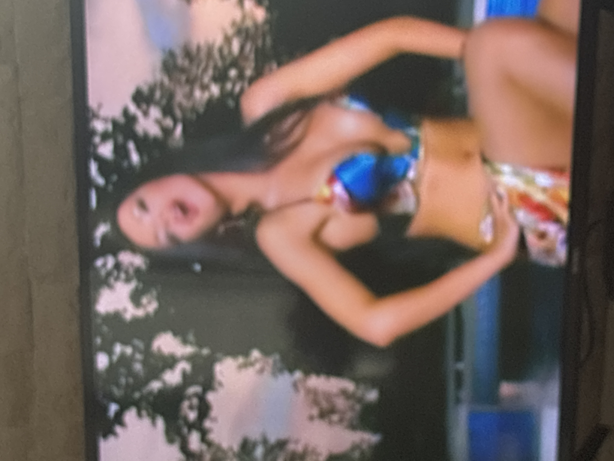 The image, rotated 90 degrees clockwise, depicts a young woman posing in an outdoor setting. She has long, dark hair and fair skin, and is wearing a vibrant, multi-colored bikini. The bikini top features patterns in blue, yellow, red, and green, with matching patterned bottoms. The woman is lying on her back, with her head turned slightly towards the viewer and her mouth slightly open, conveying a playful or expressive look. Her arms are bent, with her hands near her mid-section, and her knees are bent, lifting her legs.
The background primarily consists of lush green foliage, suggesting an outdoor natural environment such as trees or bushes. On the right side of the frame, a blue and white structure, possibly part of a building or a fence, is partially visible. The scene is brightly lit, indicating it is daytime. The image quality is somewhat blurry and grainy, characteristic of a screen capture or a low-resolution source. There is no visible text within the image.