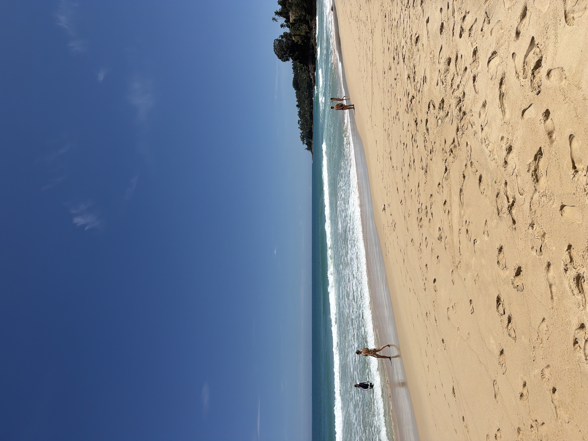 The image depicts a wide, sandy beach in Choeng Thale, Thailand, under a clear blue sky with a few wispy clouds. Gentle waves roll onto the shore, creating a foamy white surf. Several people are present on the beach and in the water. On the left side of the image, a person wearing a black and white top stands in the shallow water, facing away from the camera. To their right and slightly further up the beach, a person in a patterned bikini walks away from the water. In the distance, on the right side of the frame, two figures are walking along the shoreline. The beach is marked with numerous footprints in the sand. Lush green vegetation and trees cover a hillside in the background, with a hint of palm trees visible. The sea stretches out to the horizon, appearing calm and turquoise.