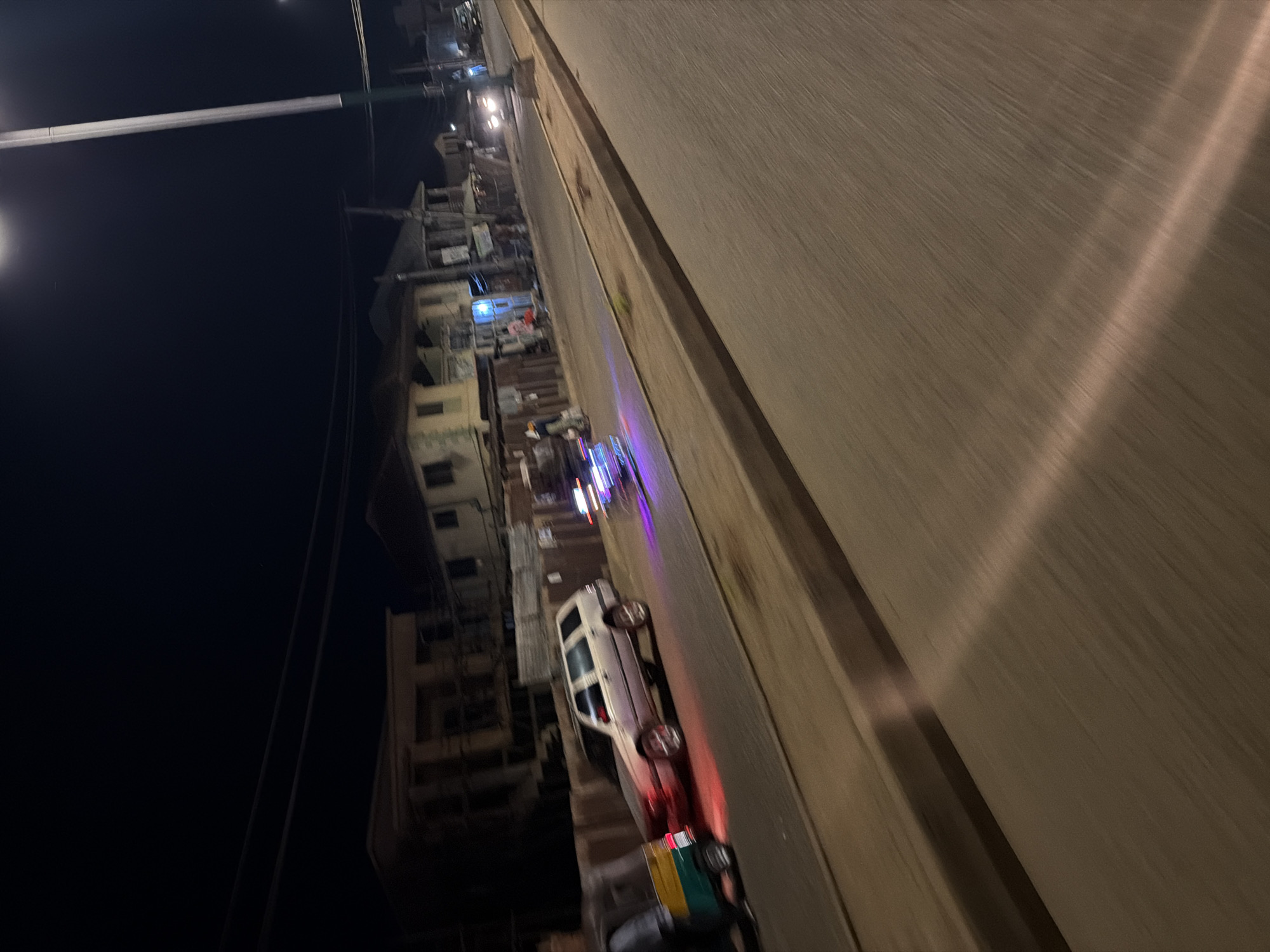 A nighttime street scene in Bukuru, Nigeria, features a street with passing vehicles and buildings lining the side. A light-colored station wagon is parked on the left side of the road, with its red brake lights illuminated. To its left, a three-wheeled auto-rickshaw is partially visible. Further down the road, other vehicles are moving, their headlights and taillights creating streaks of light due to motion blur. Buildings, appearing to be residential or commercial, with visible windows and some illuminated interiors, are situated along the street. A tall street lamp illuminates a portion of the scene. The overall impression is of a busy street at night in an urban environment.