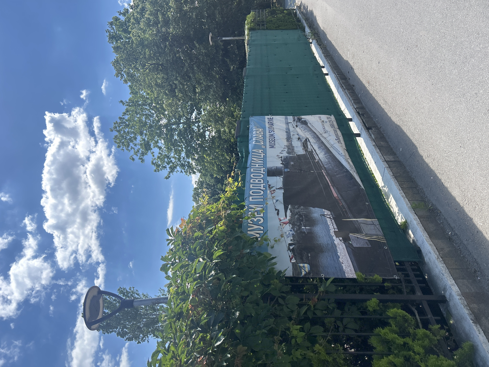 The image shows a banner for the "Museum Submarine" in Varna, Bulgaria. The banner features an image of a submarine and is written in Cyrillic and English. The setting appears to be outdoors, with lush green trees and a bright blue sky with fluffy white clouds. The sun is shining, suggesting it is daytime. The banner is partially obscured by green foliage and a metal fence. A grey asphalt path runs alongside the fence. The overall impression is a sunny day at an outdoor attraction, possibly on the coast given the submarine theme. No people are visible in the image.