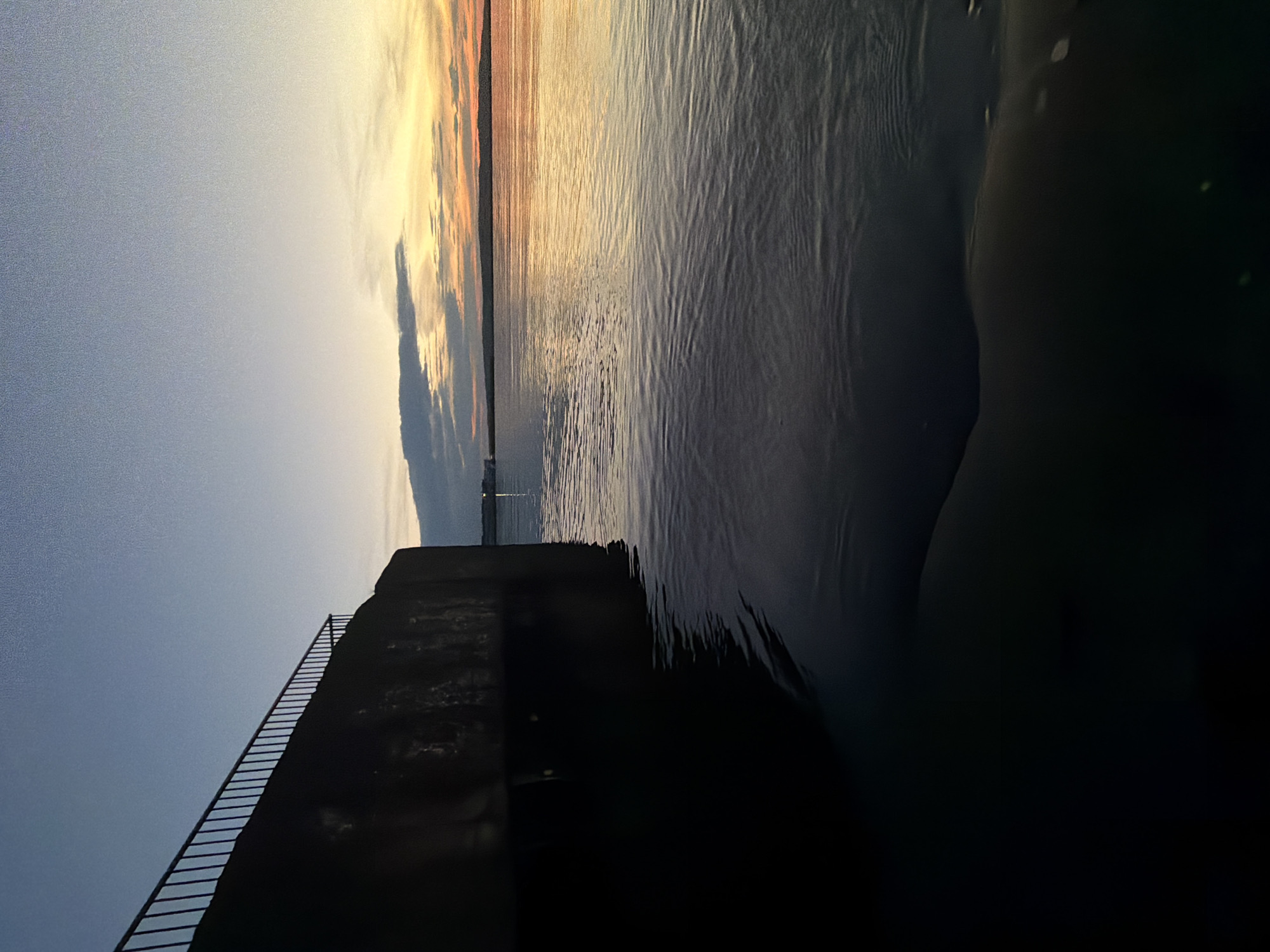 The image shows a dark, silhouetted structure with a railing running along its edge, possibly a pier or seawall. The structure leads into a body of water that reflects the sky, which is filled with soft, warm colors of sunset or sunrise, indicating either dawn or dusk. The sky transitions from a clear, pale blue on the left to warmer hues of orange and yellow where the sun is likely setting or rising, with some dark clouds adding depth. The water has a textured surface, with ripples catching the light. In the distance, a thin line of land or a structure is visible on the horizon, and a faint light can be seen in the distance. The overall mood is peaceful and serene, evoked by the soft light and the stillness of the water. The scene appears to be an outdoor setting at a waterside location. There is no visible text in the image.