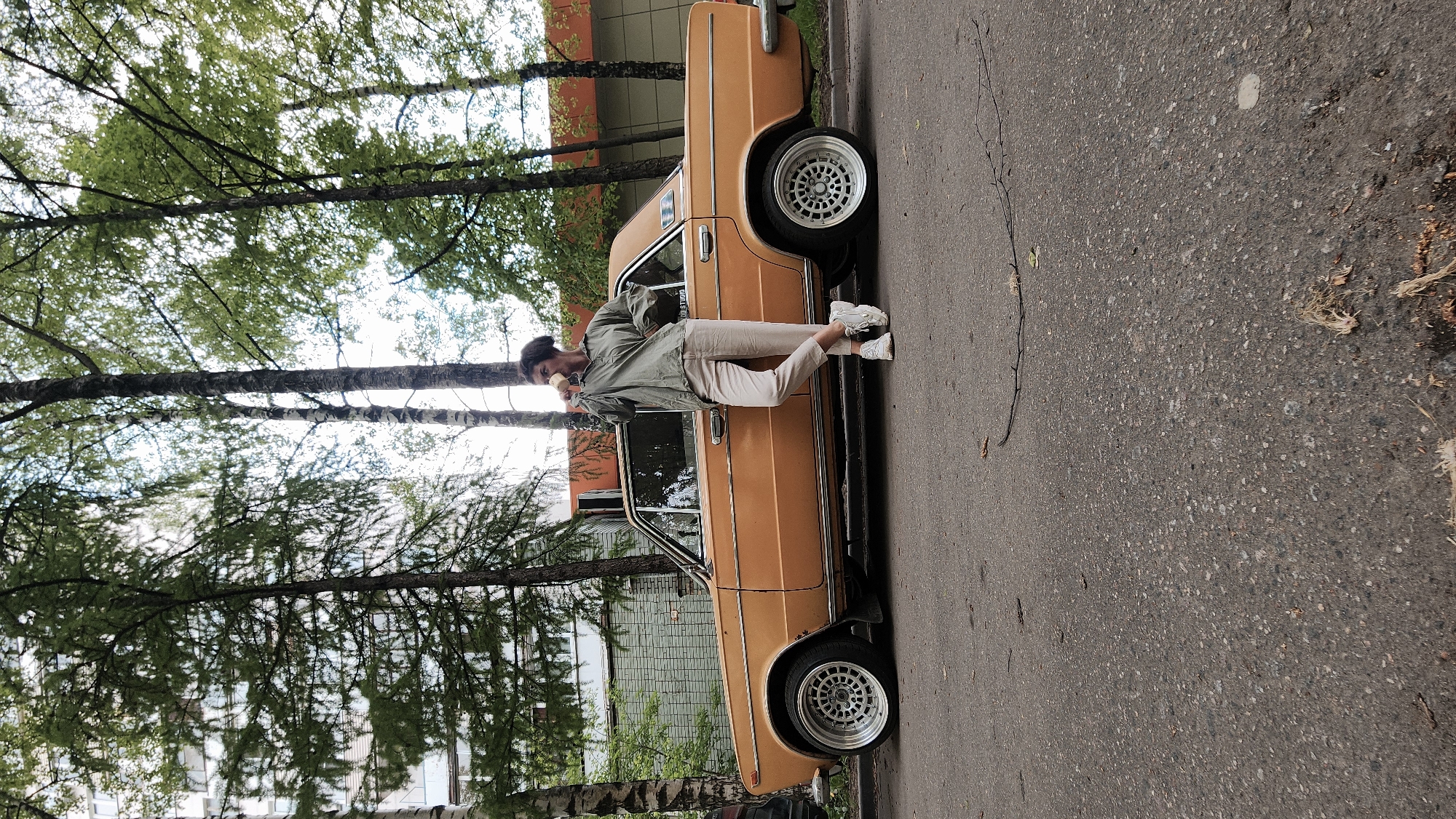 A woman leans against an orange vintage car parked on an asphalt road. She is wearing an olive green jacket, beige pants, and white sneakers, and appears to be holding a cup or mug in her hand, possibly drinking from it. The car is parked on the side of the road with trees in the background. Buildings can be seen in the distance, suggesting an urban environment. Based on the architectural style of the buildings and the overall ambiance, the location is possibly in Moscow, Russia.